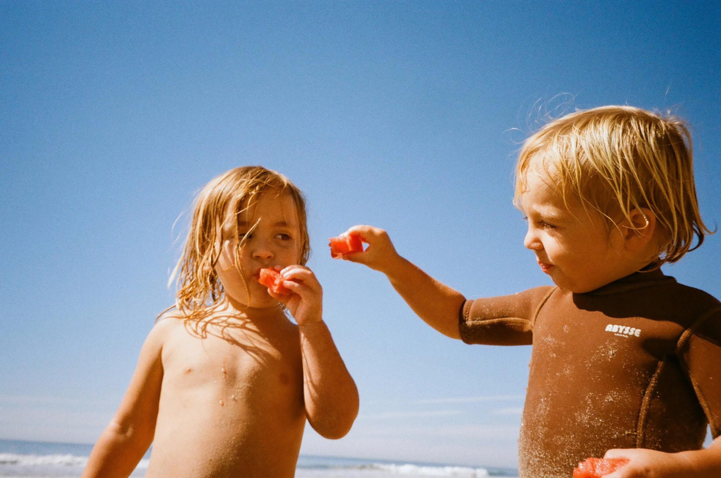 Two young children with sandy skin and blonde hair enjoying watermelon on the beach under a clear blue sky.