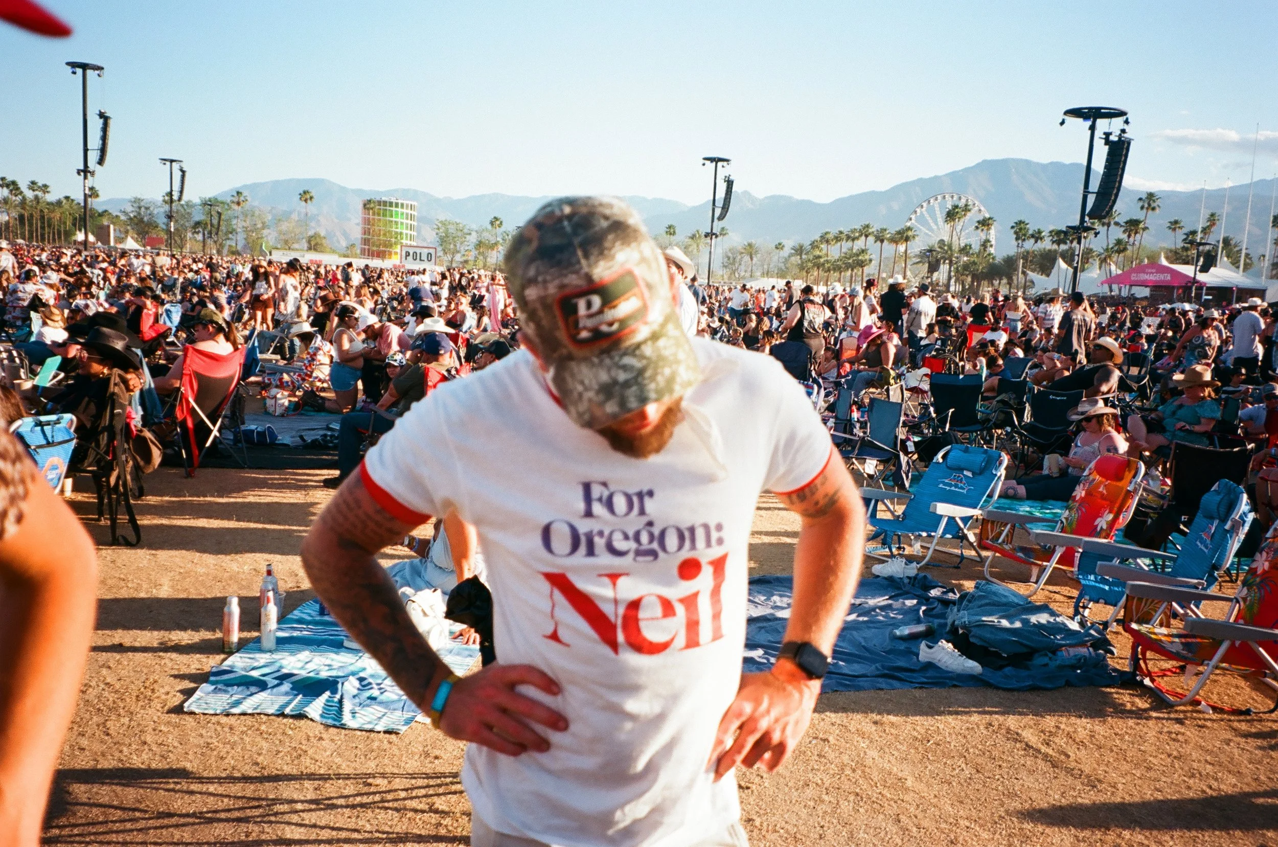 A man wearing a white t-shirt with red text that says 'For Oregon: Neil' and a camouflage baseball cap, standing with hands on hips at a crowded outdoor festival or concert, with many people sitting on chairs in the background and mountains in the distance.