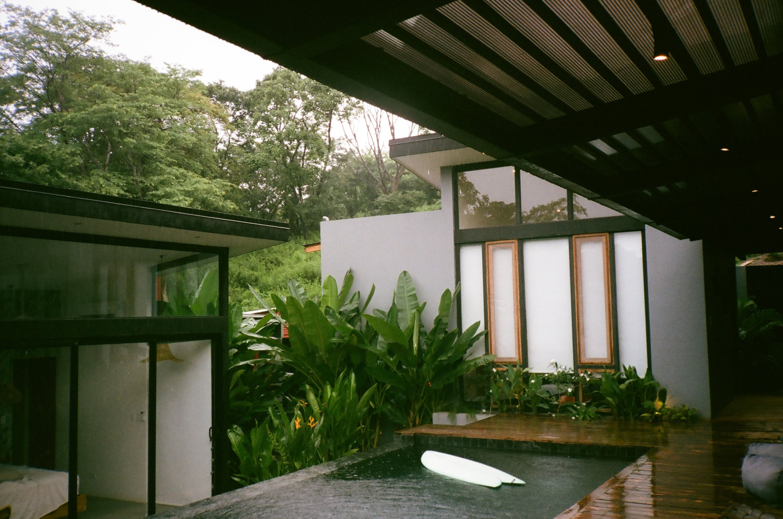 Modern house with white walls, black accents, large windows, surrounded by lush green trees and plants, with a wooden deck and outdoor seating area under an overhang.