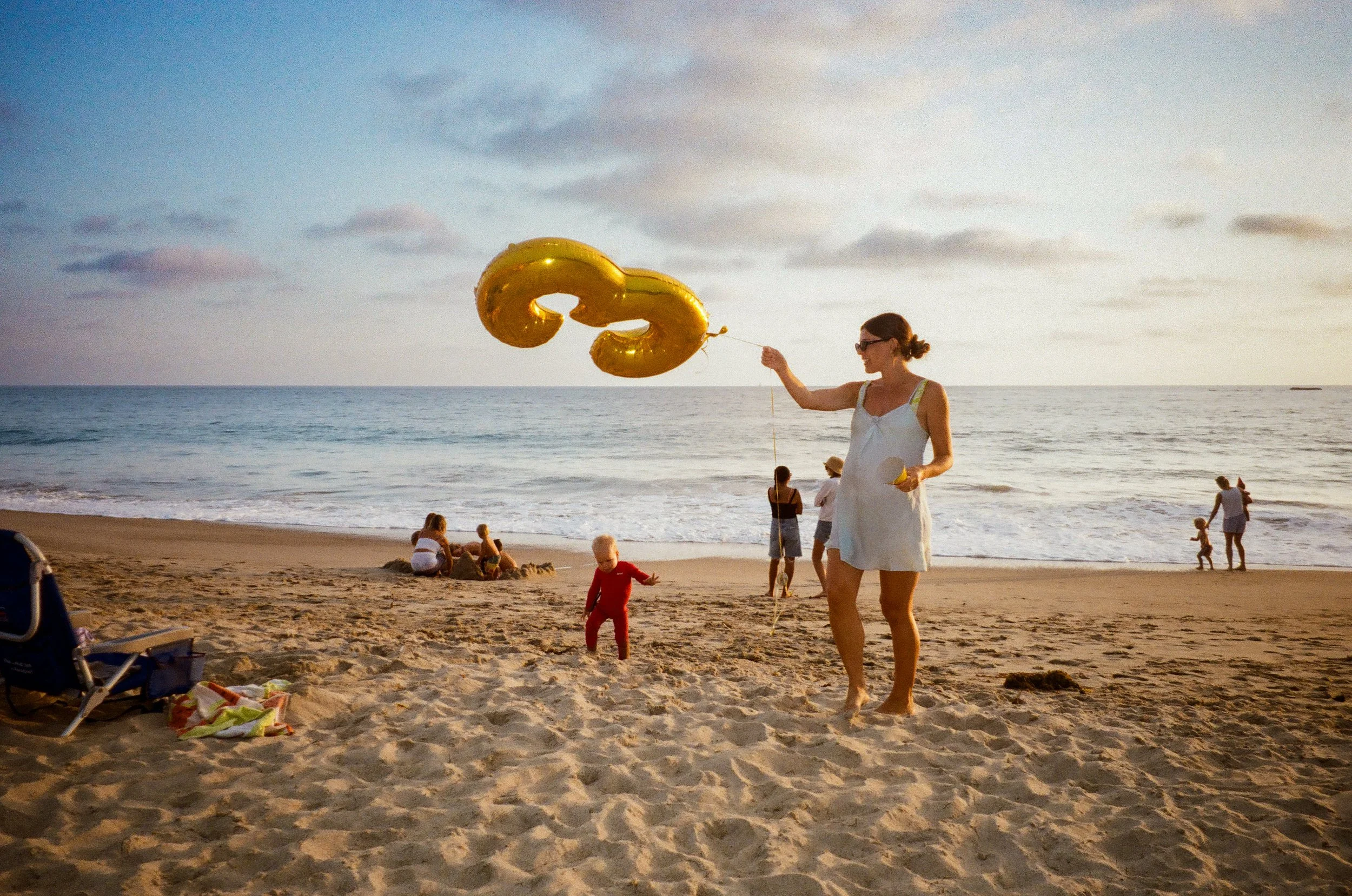 A woman on a beach holding two gold balloons shaped like the number 30. Other people are relaxing and walking along the shoreline during sunset.