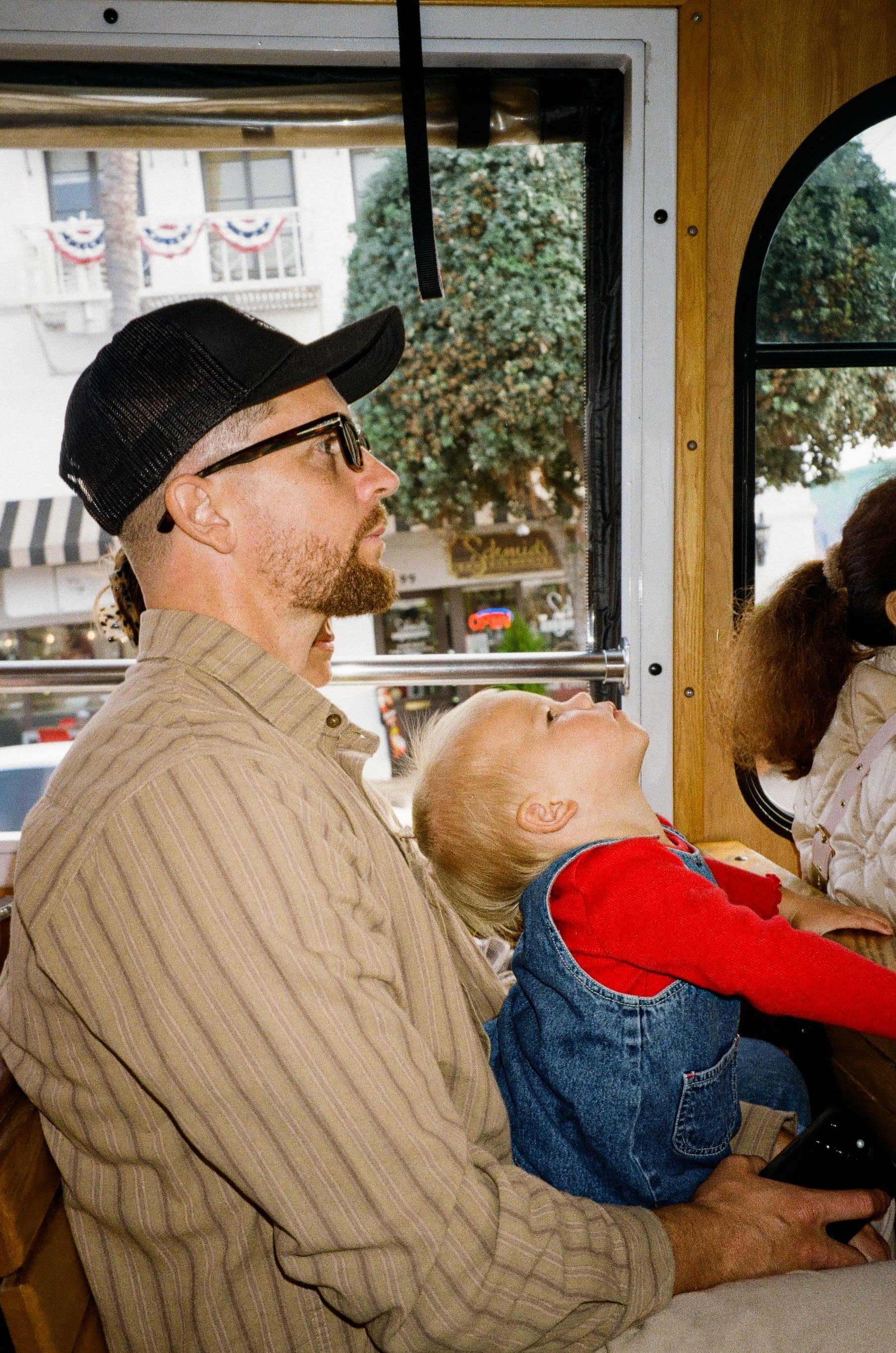 A man wearing glasses, a black cap, and a beige striped shirt sitting next to a young blonde girl in a red shirt and denim overalls. They are on a bus, with a window showing a street scene behind them.