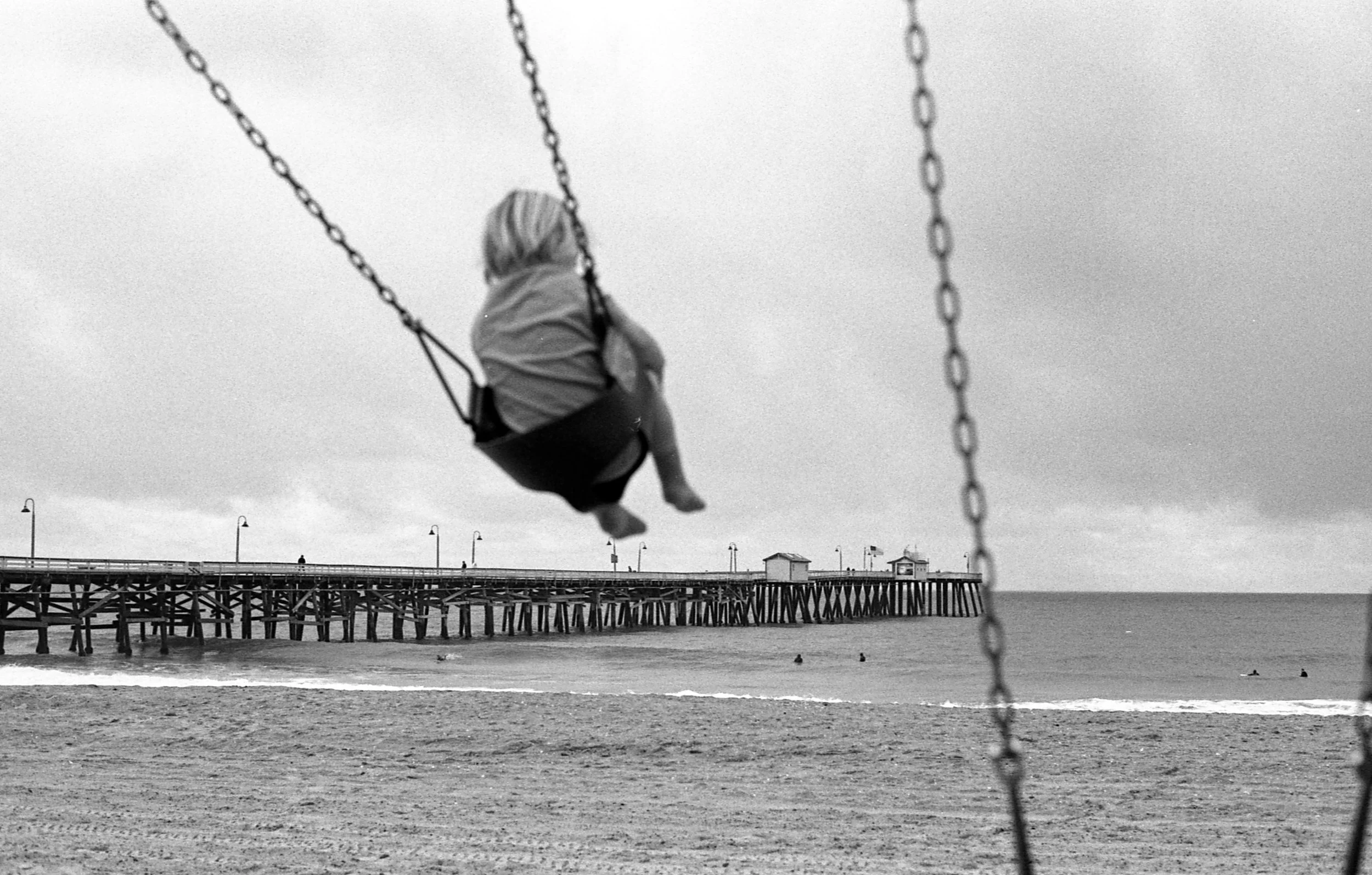 A child on a swing at the beach with a pier in the background, grayscale photograph.