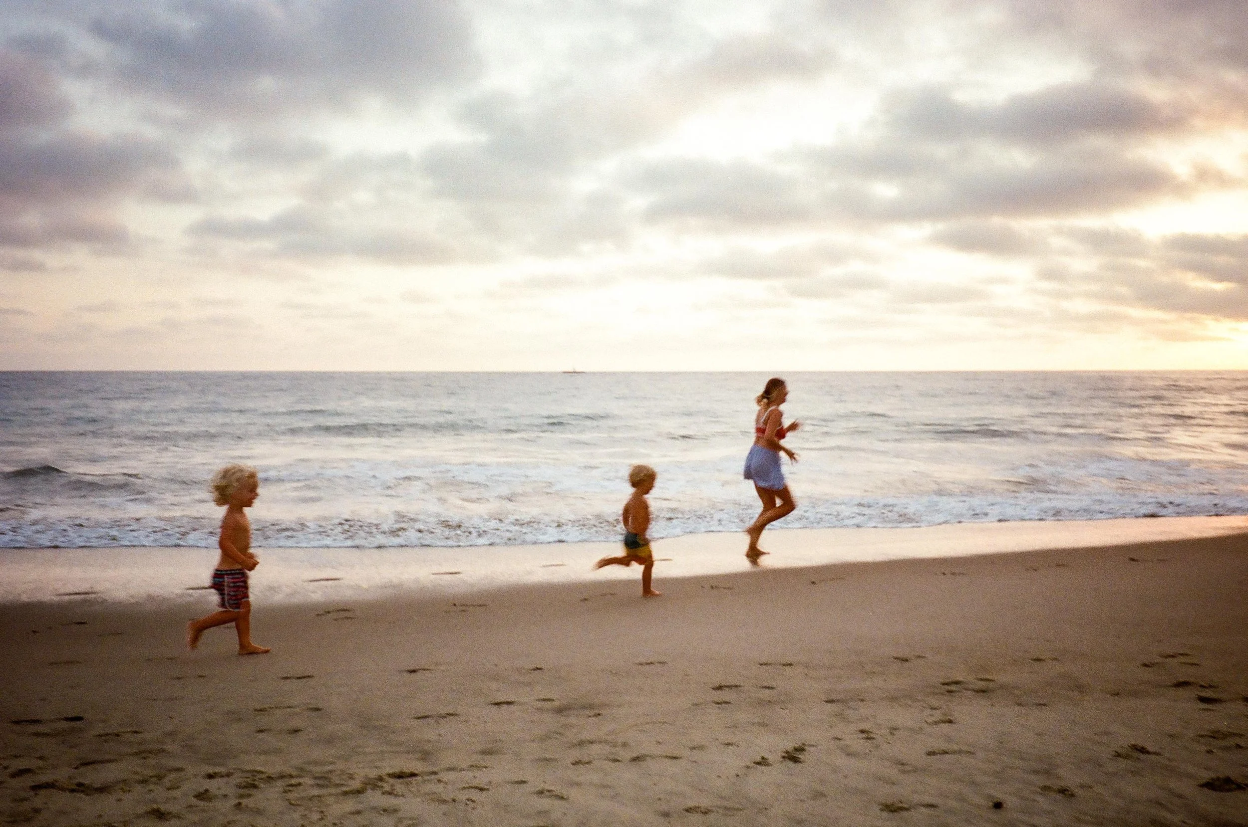 A woman and three children running along a sandy beach at sunset, with the ocean and cloudy sky in the background.