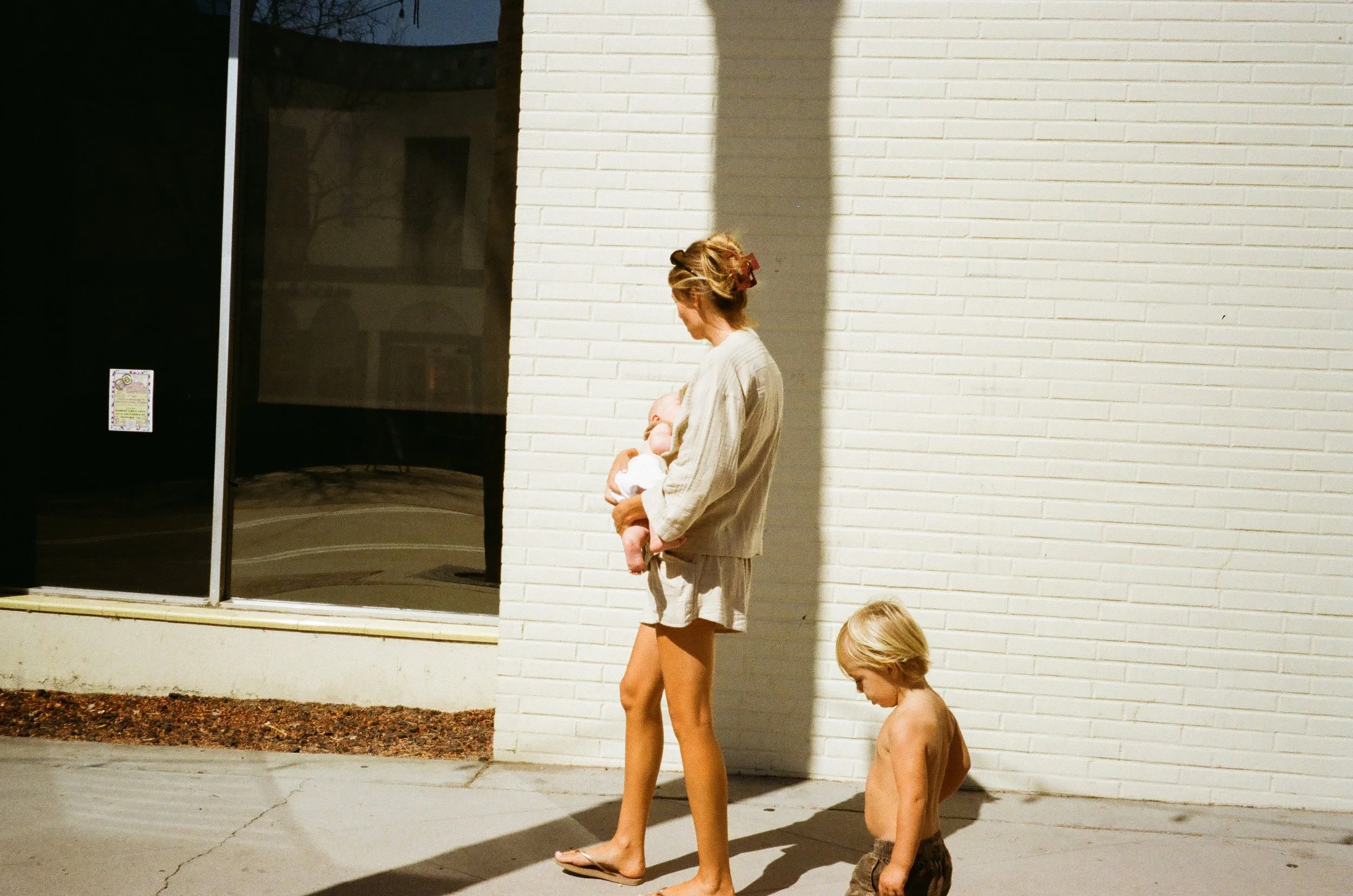 A woman holding a baby and a young boy walking on a sidewalk outside near a white brick building.