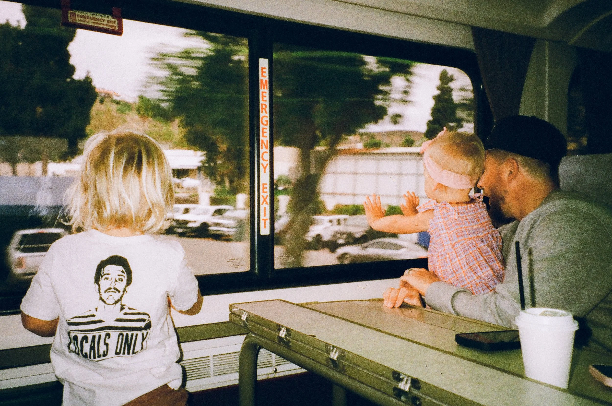 A man with a beard and a black baseball cap, a young girl with a pink headband, and a child with blonde hair looking out a bus window. The girl is reaching toward the window while the man is smiling. The child's back shows a white T-shirt with a graphic of a man's face and the words 'Locals Only'. A white cup with a straw and a phone are on the table in front of them. Outside the window, there are trees and parked cars.