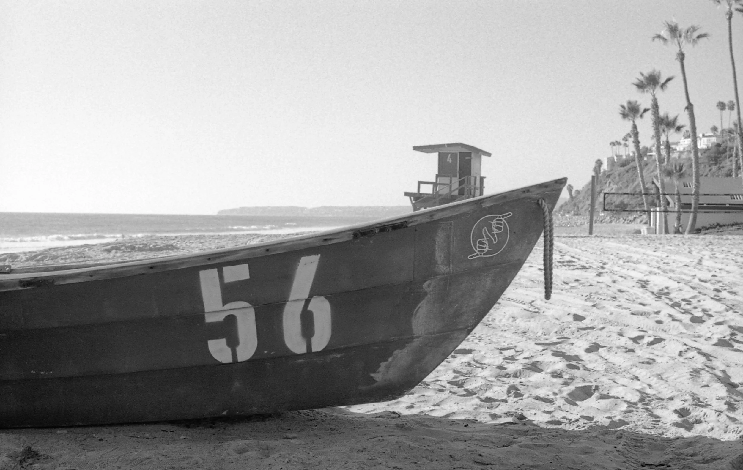 A lifeboat with the number 56 on it on a sandy beach, with a lifeguard tower, beach chairs, and palm trees in the background.