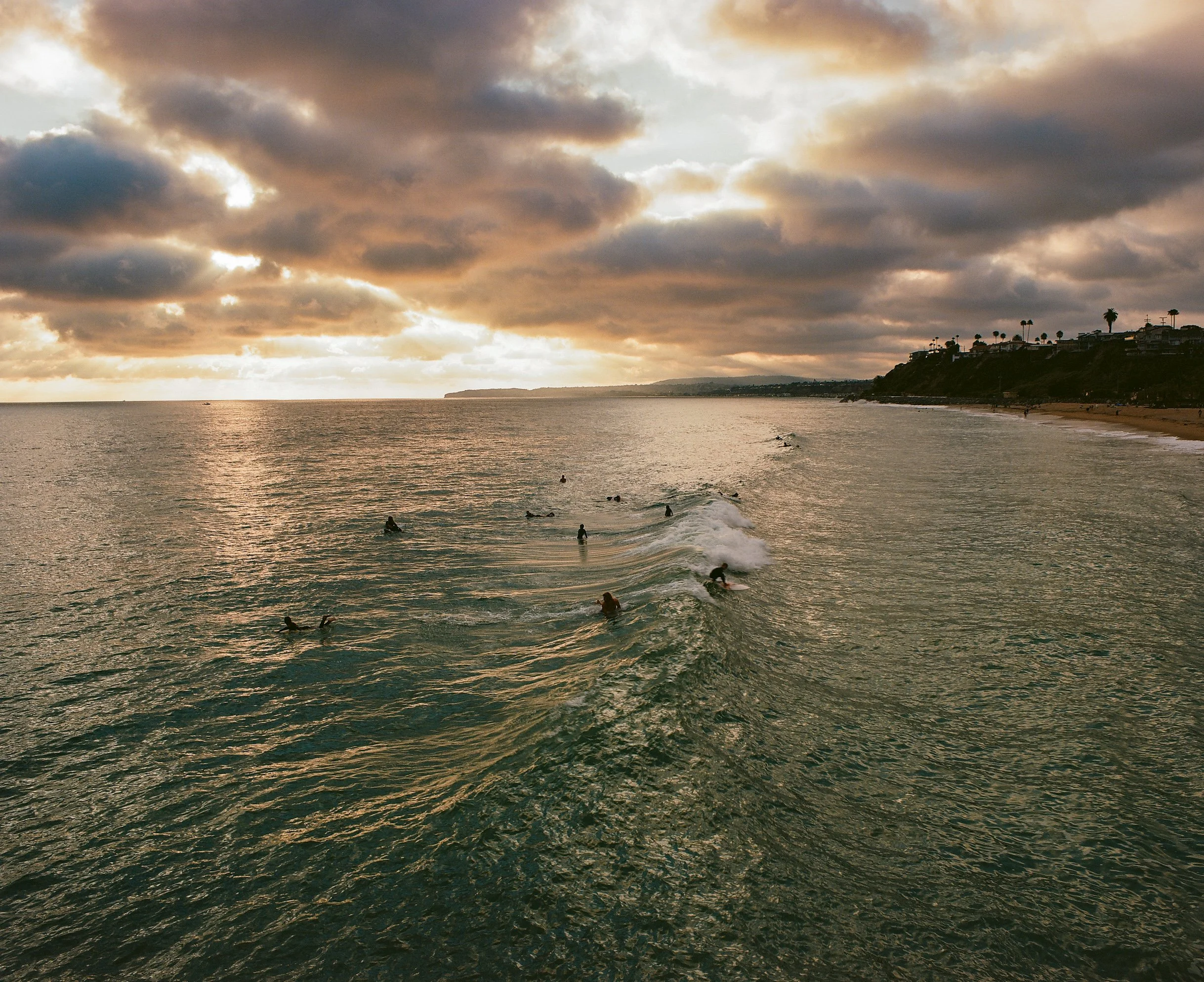 Sunset over the ocean with many surfers in the water, some riding waves and others waiting, and a shoreline with trees and houses in the distance.