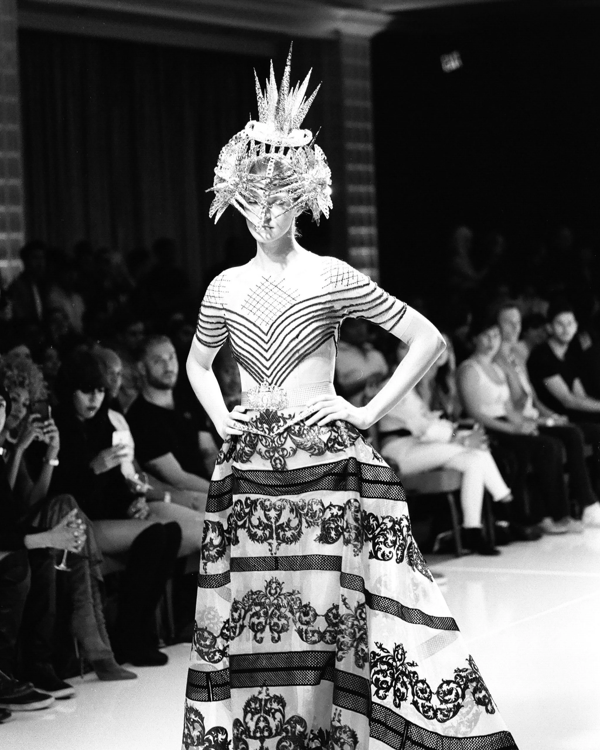 A fashion model walking on runway during a fashion show, wearing a large ornate headpiece and a detailed dress with intricate patterns, while audience members watch.