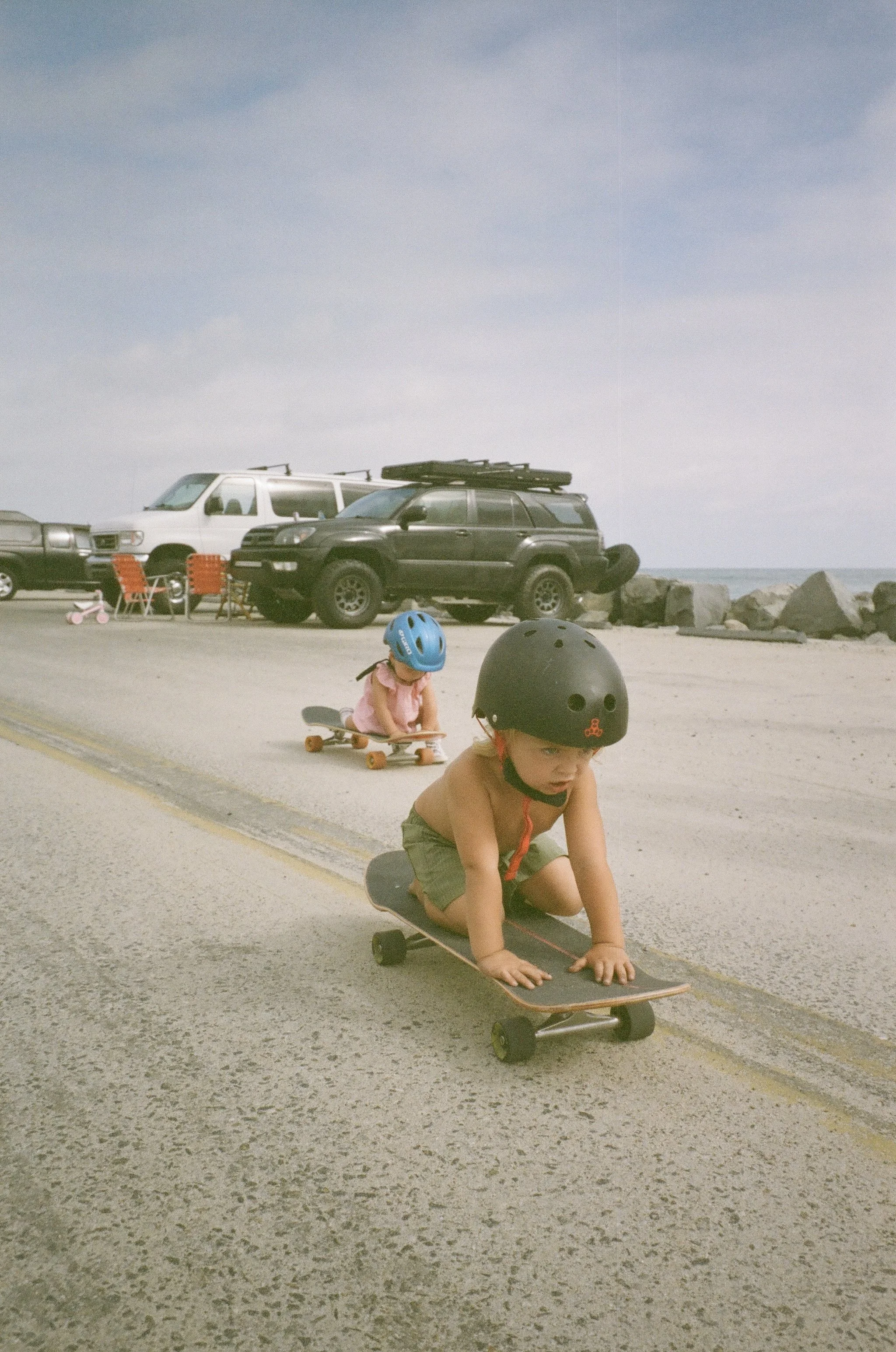 Kids riding skateboards on the beach road, wearing helmets, with parked cars and rocks in the background.