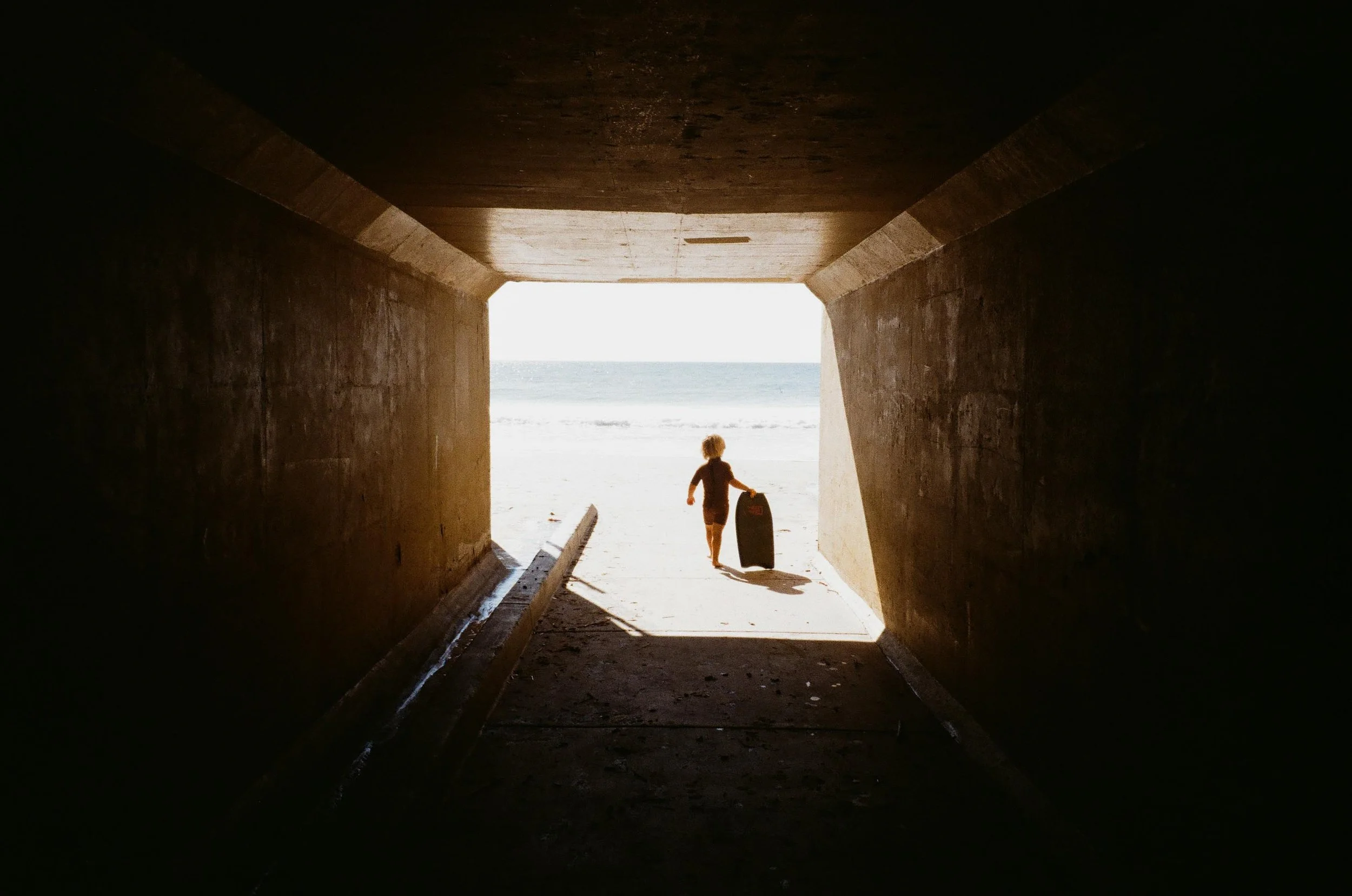 A child carrying a suitcase walking through a tunnel towards the beach and ocean.