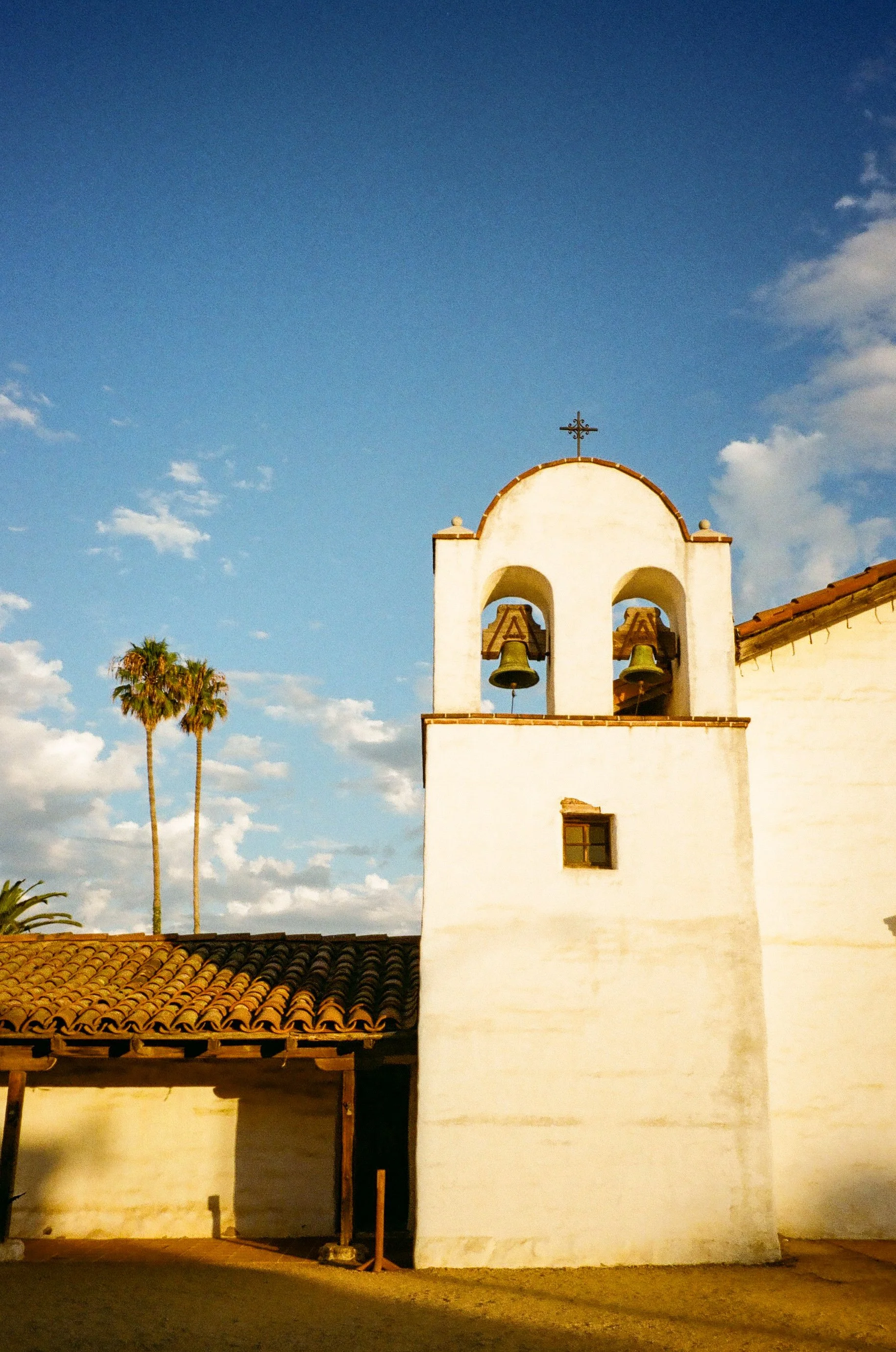 A small white church with a bell tower and cross on top, two bells, and palm trees against a blue sky with scattered clouds.