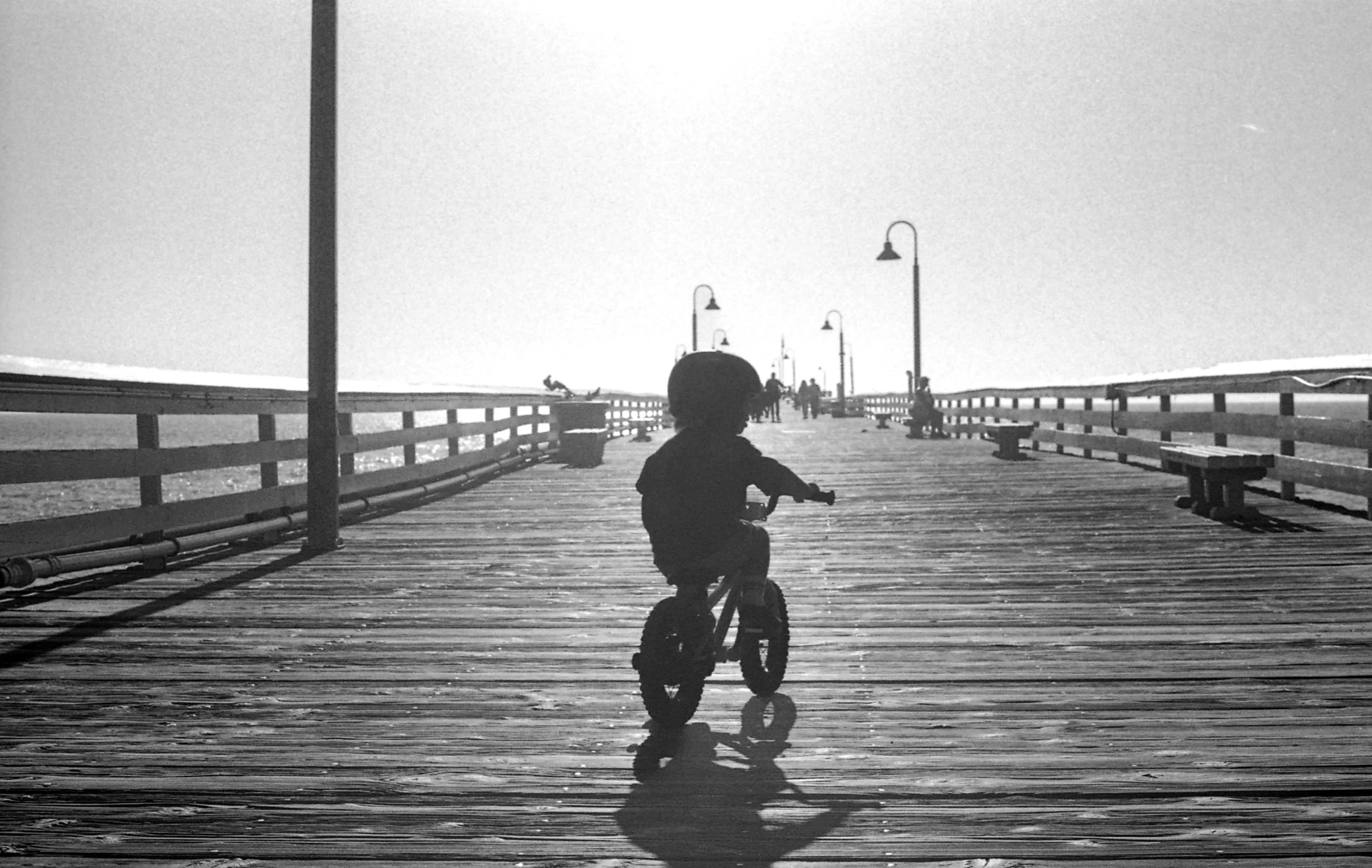 Child riding a bicycle on a wooden pier with lampposts and benches, in black and white.