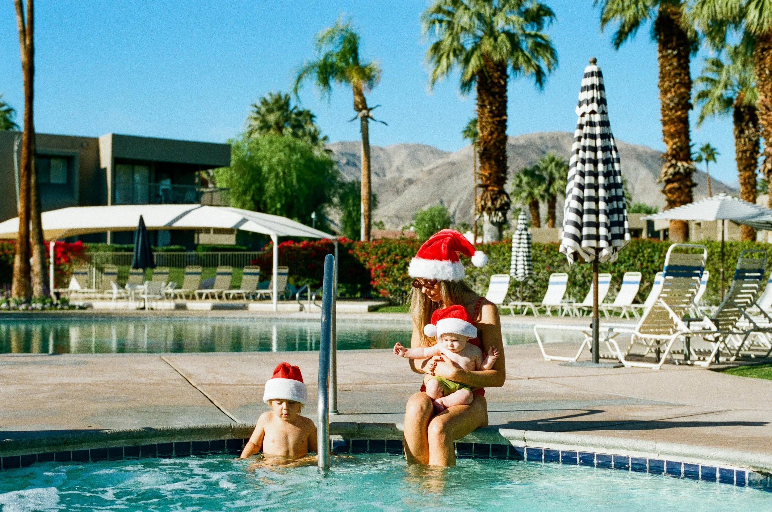 A woman and two children wearing Santa hats at a poolside during daytime. One child is in the water, the woman is sitting on the pool edge holding the other child on her lap. The background shows lounge chairs, umbrellas, palm trees, and distant mountains.