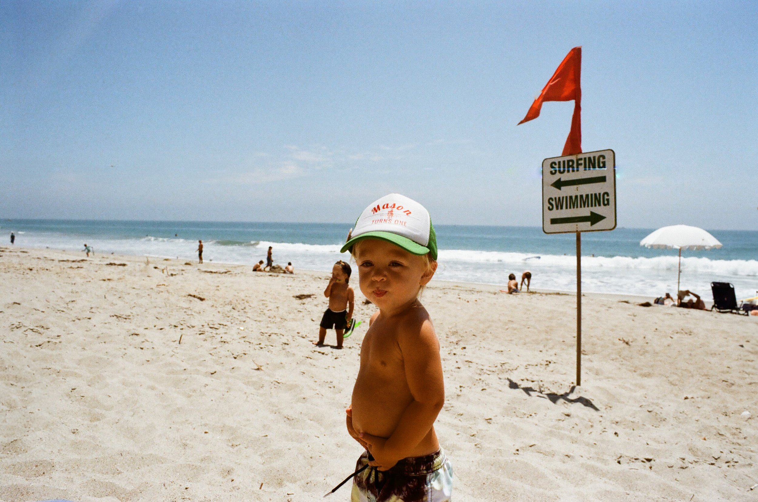 A young boy in swim trunks and a white cap with green trim stands on a sandy beach near the water, with a sign indicating swimming and surfing areas and a red flag behind him.