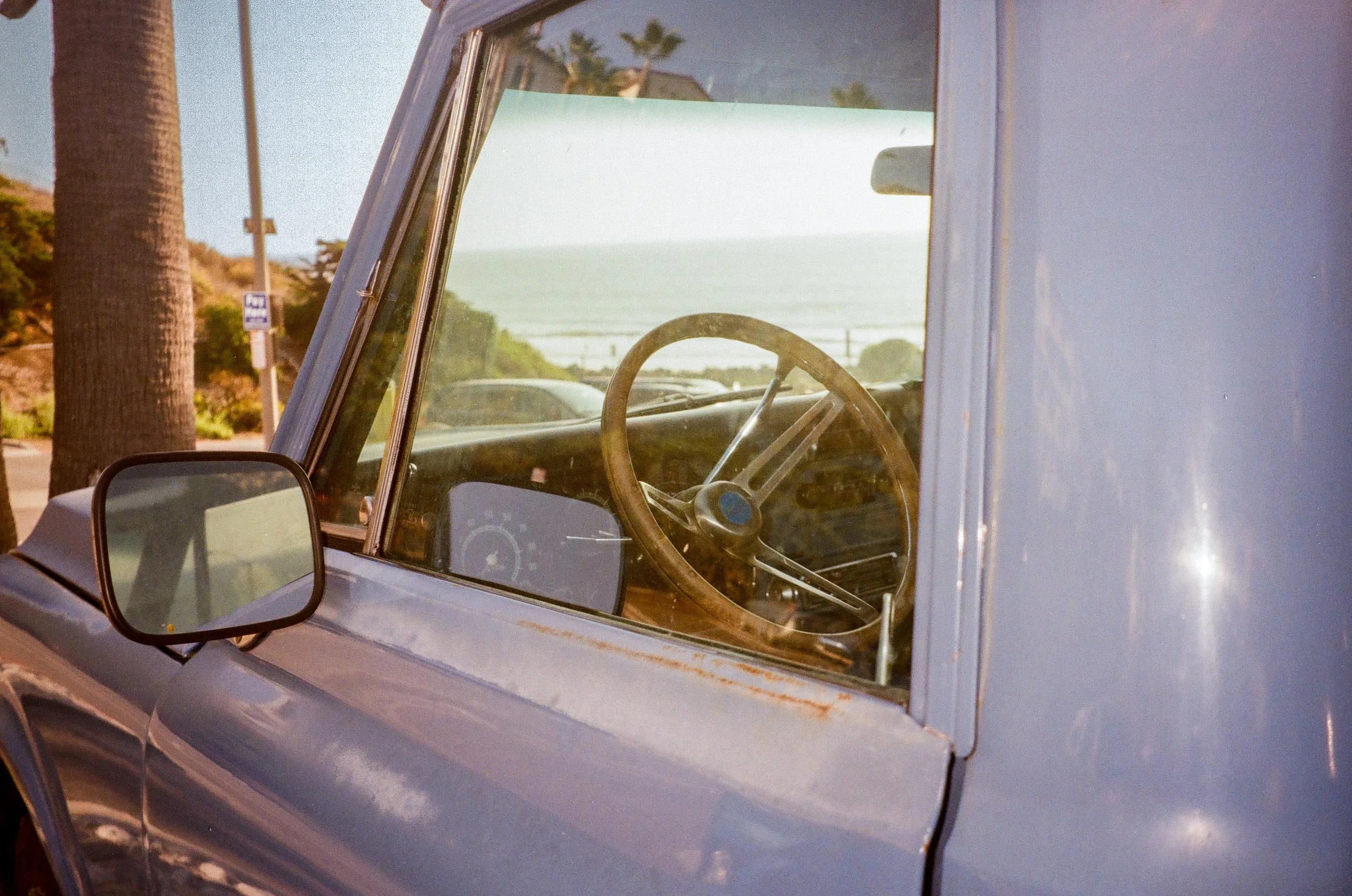 Close-up view of the front windshield and interior of a vintage car parked outdoors near palm trees, with a scenic view of the ocean and blue sky in the background.