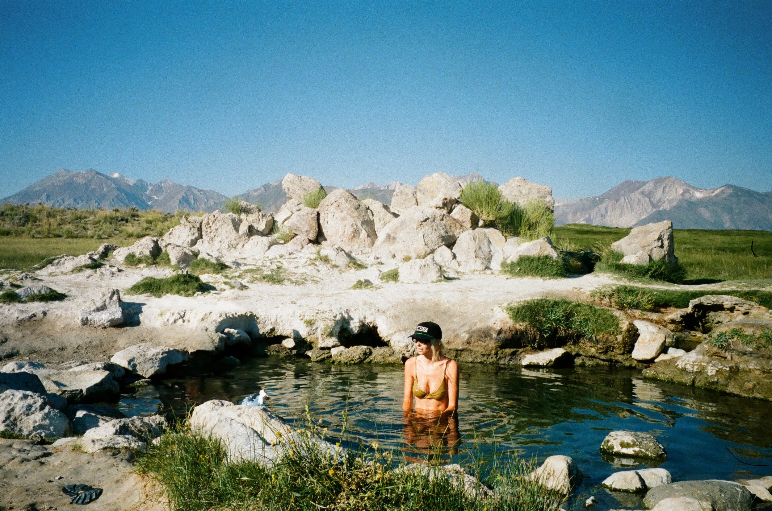A woman in a bikini and cap standing in a small natural hot spring surrounded by rocks, with mountains in the background under a clear blue sky.