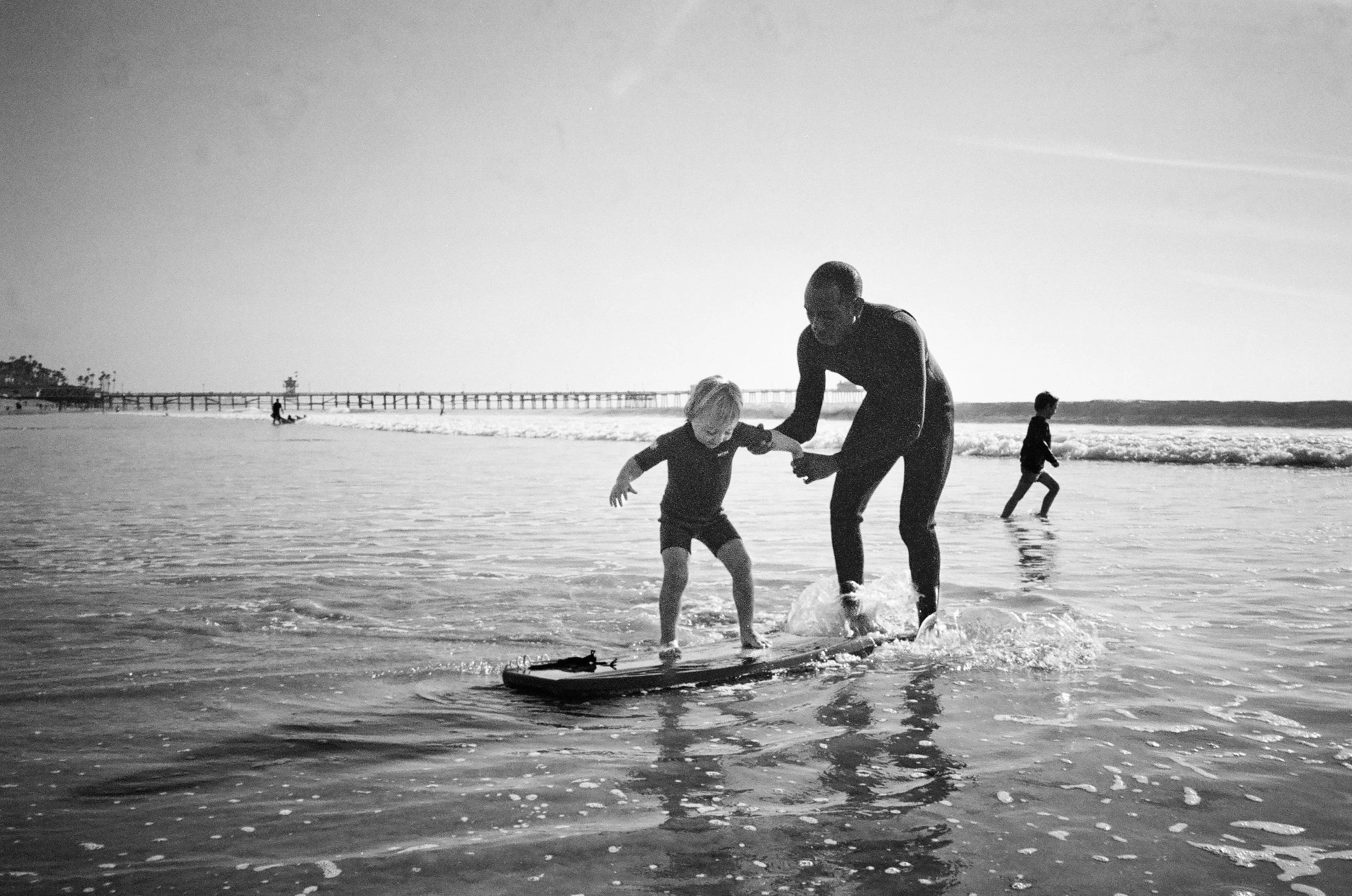 A man and a young child riding a skateboard in the shallow ocean water, with other children playing in the background near the shoreline and a pier extending into the ocean.