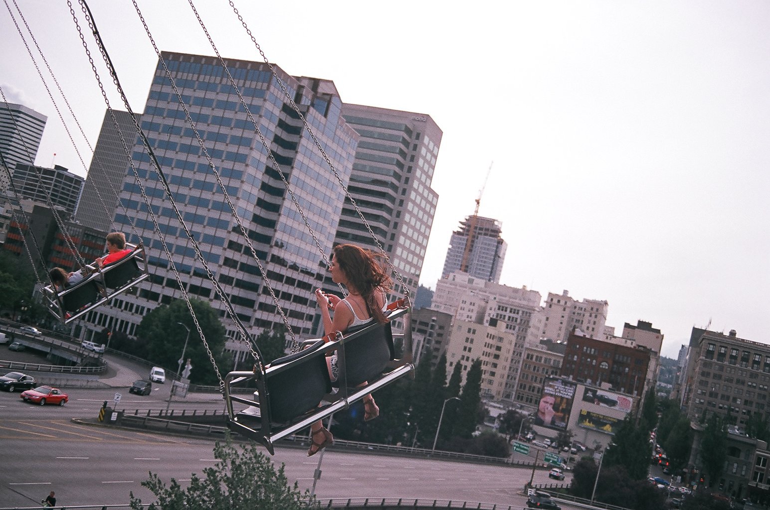 A woman and a child on a swing ride above a city street with skyscrapers in the background.