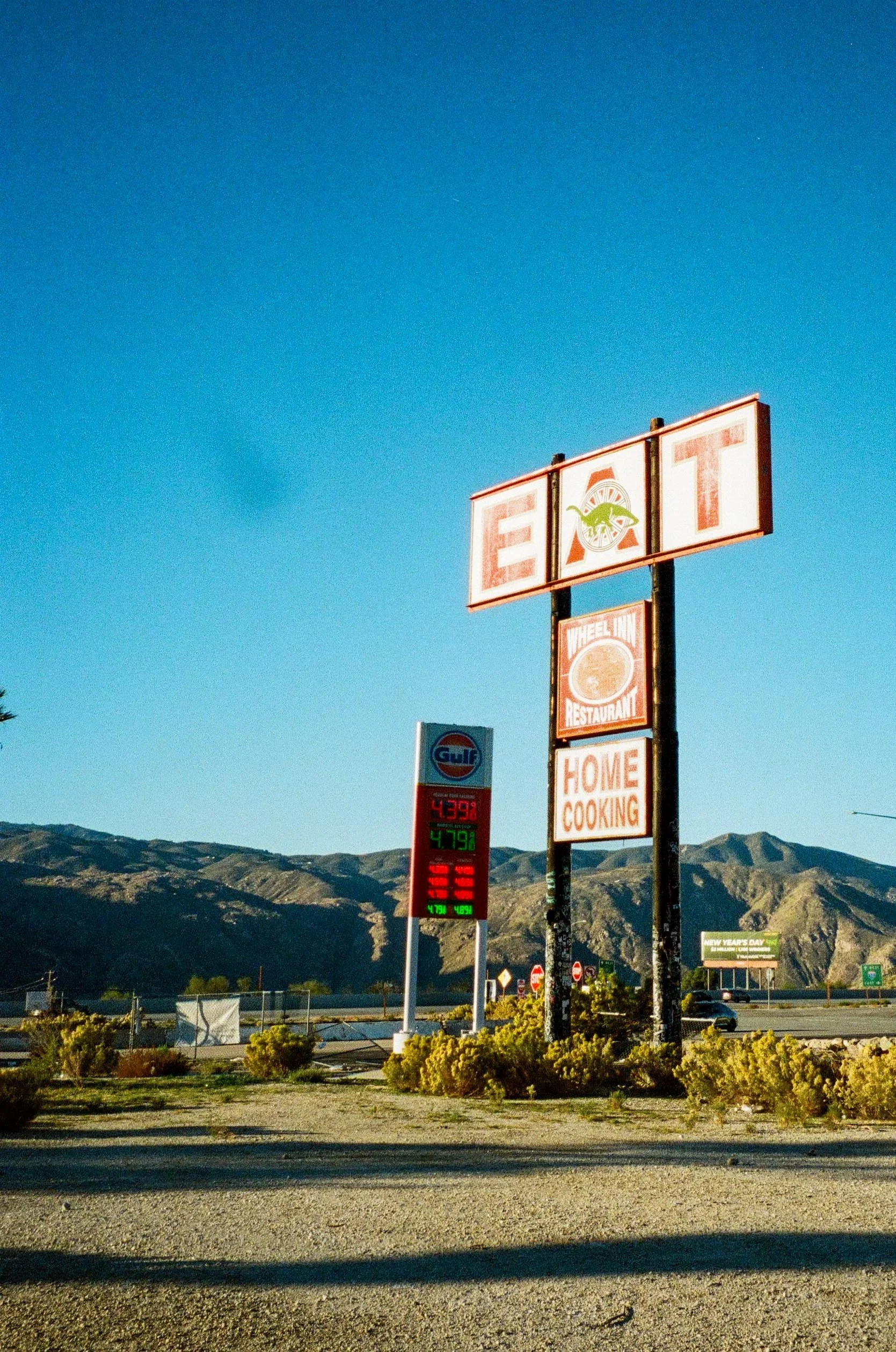A sign for an eatery called EAT with a dinosaur logo, featuring an indoor restaurant and home cooking options, located near a Gulf gas station with fuel prices displayed. Mountain range in the background under a clear blue sky.