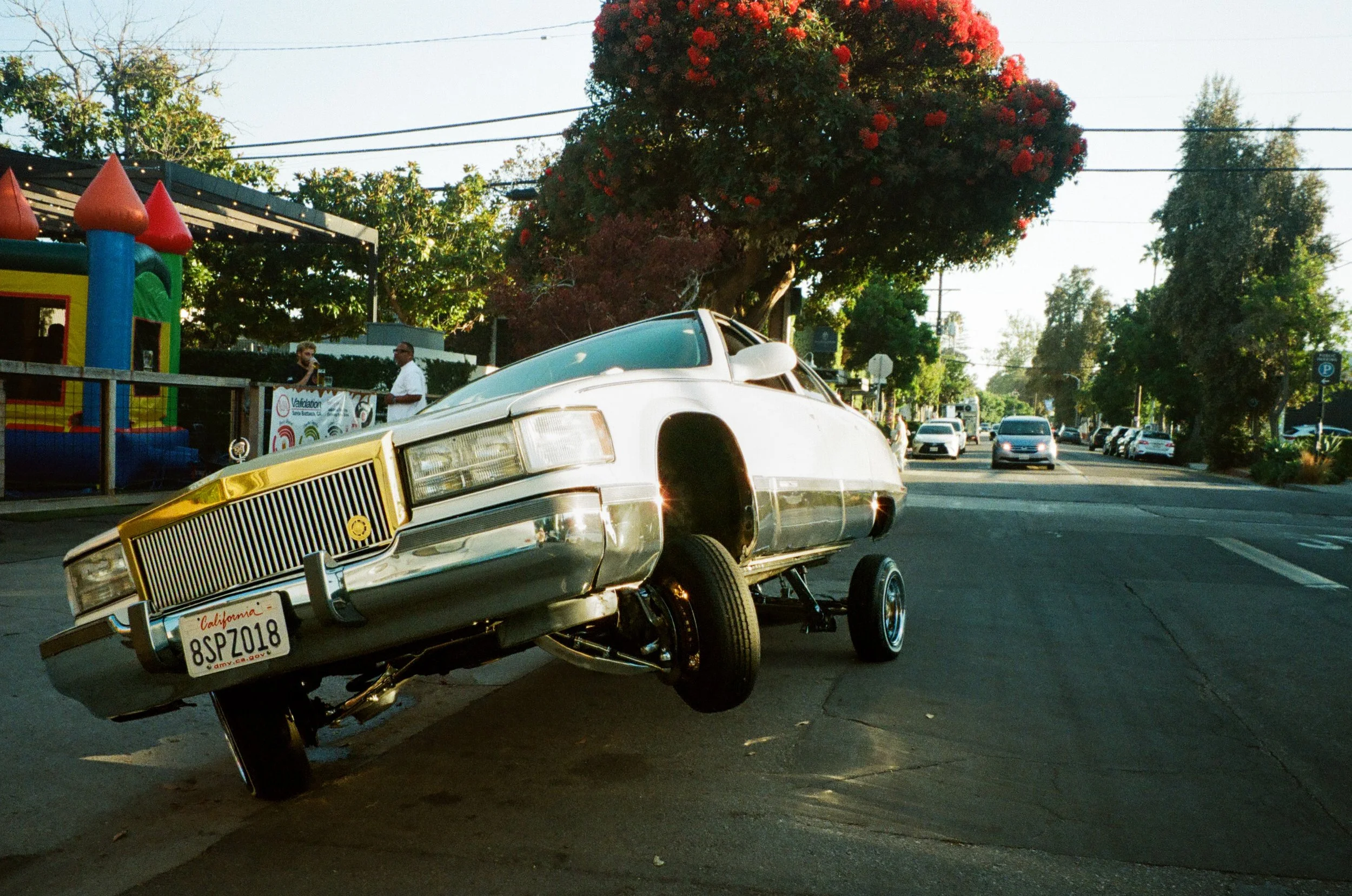 A vintage car with the front wheels elevated, balanced on two tires, on a street with trees and cars in the background. A bounce house and people are visible on the sidewalk.