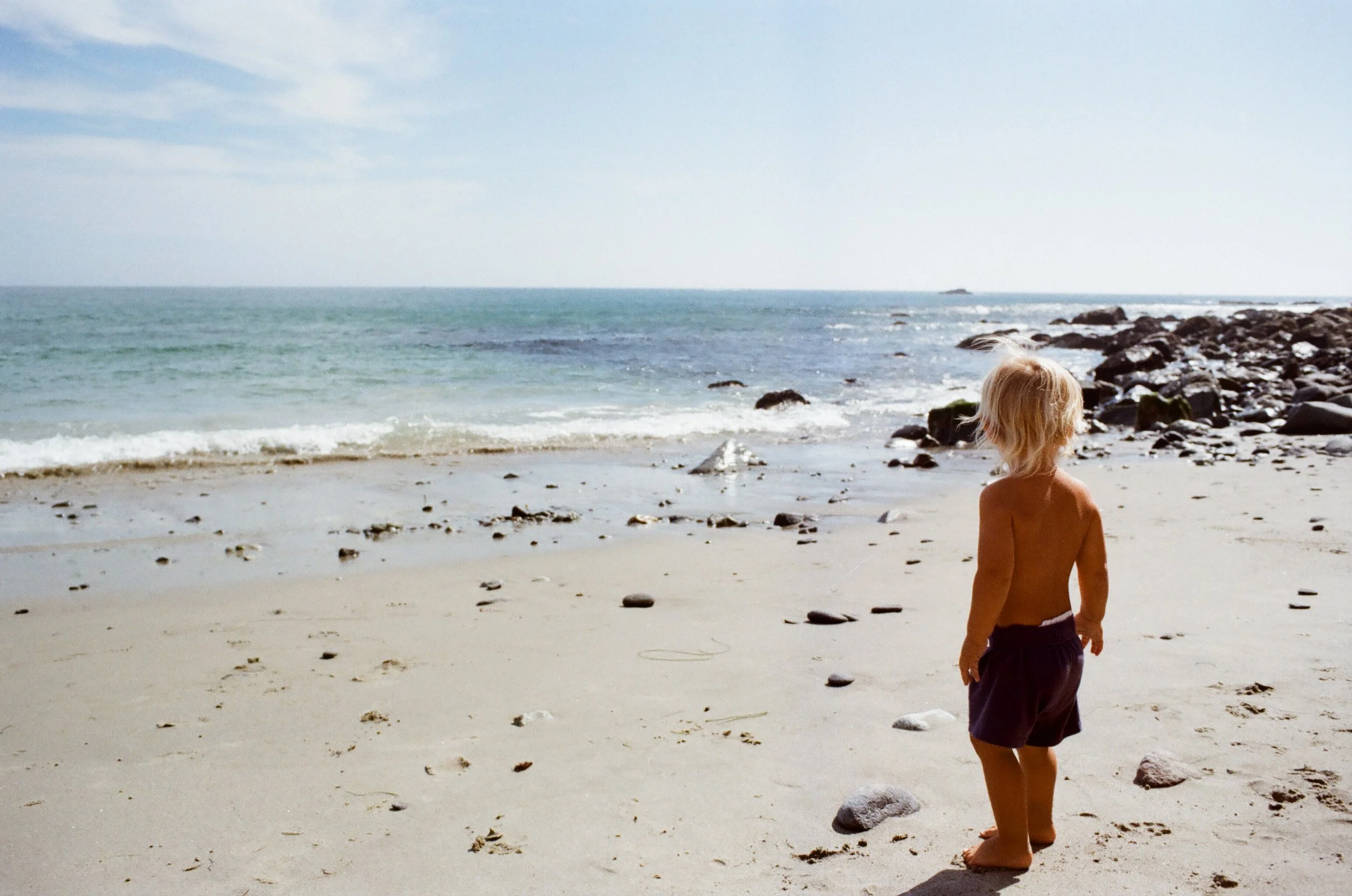 A young child with blonde hair, wearing shorts, standing on a sandy beach looking toward the ocean, with rocks along the shoreline and a blue sky overhead.
