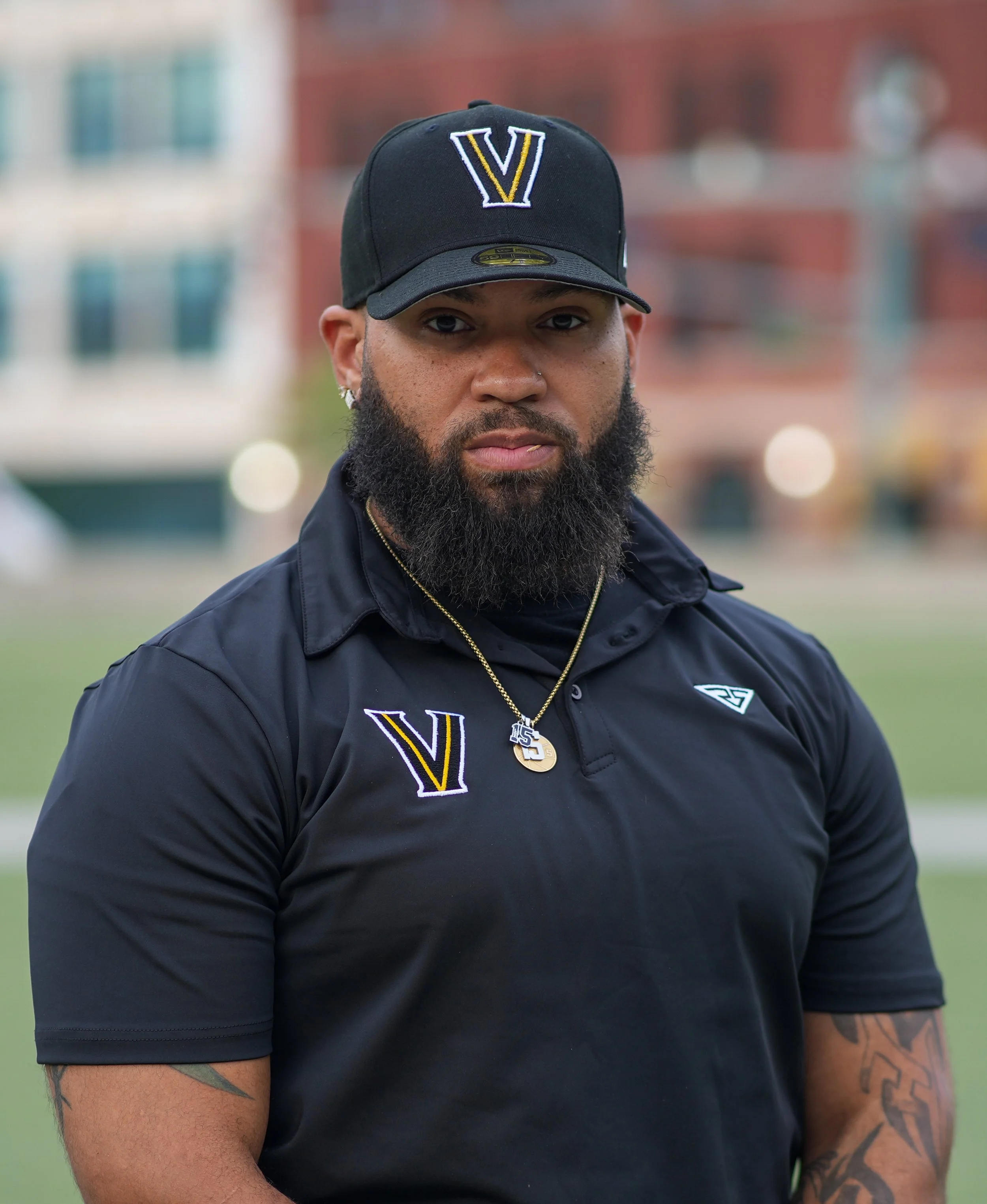 A man with a beard and tattoos wearing a black V logo cap and black sports shirt, standing outdoors with blurred buildings in the background.