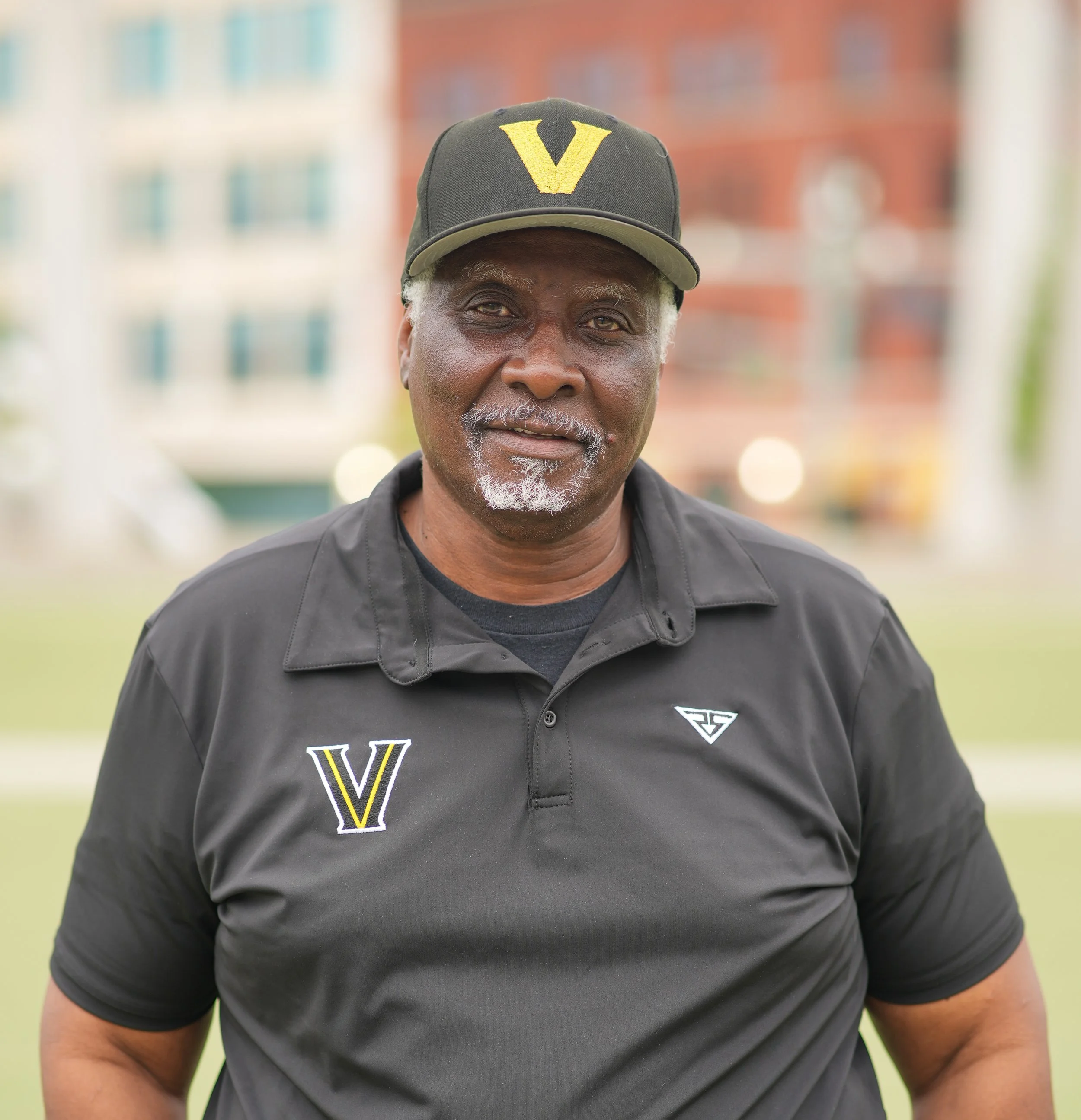 An older man with gray hair and beard wearing a black hat with a yellow 'V', a black polo shirt with a yellow 'V' and a logo badge, standing outdoors in front of a blurred cityscape.