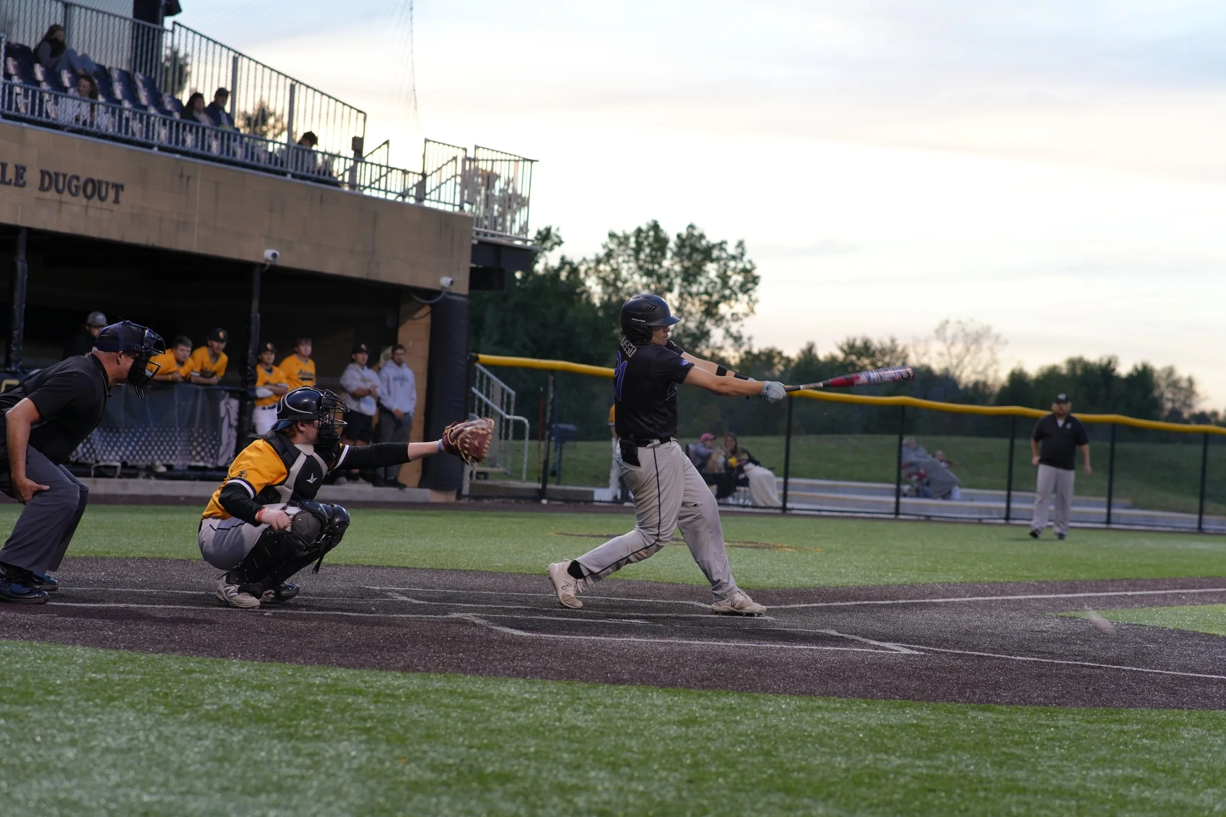 A baseball batter swinging at a pitch during a game, with the catcher and umpire crouched behind, and spectators watching from the dugout and seating area in the background.