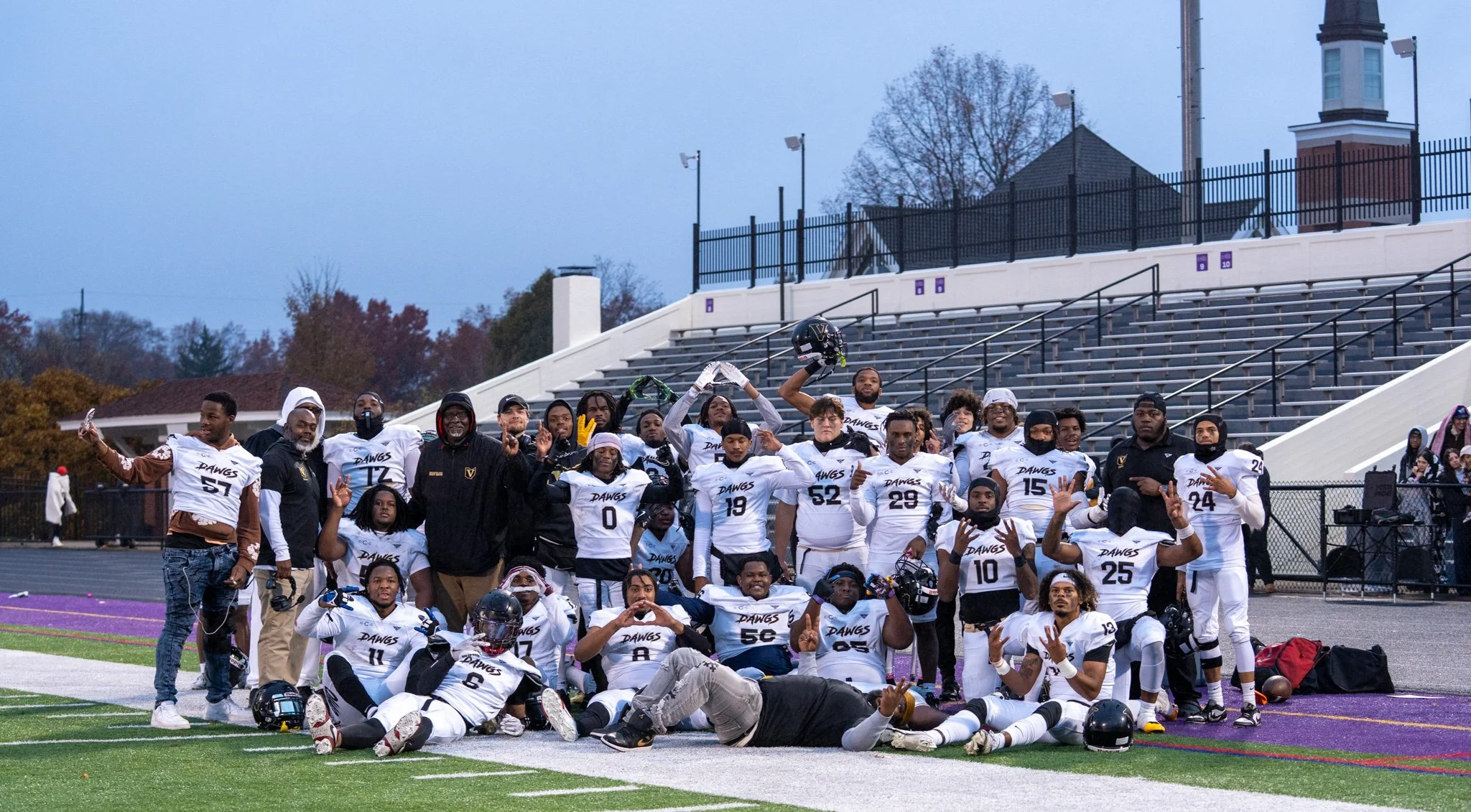 Group of football players and coaches posing on the field after a game, with some making peace signs and flexing muscles, during evening or late afternoon.