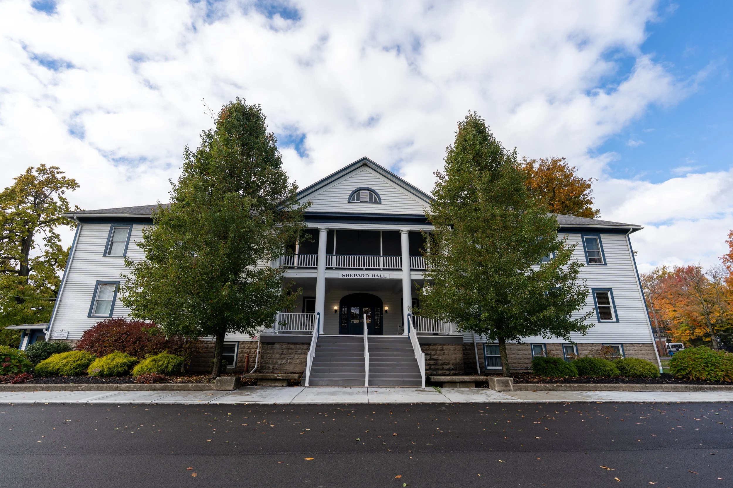 A white, three-story building with a sign that says 'Shepard Hall' above the entrance, surrounded by trees, with a sidewalk and street in the foreground and a partly cloudy sky above.