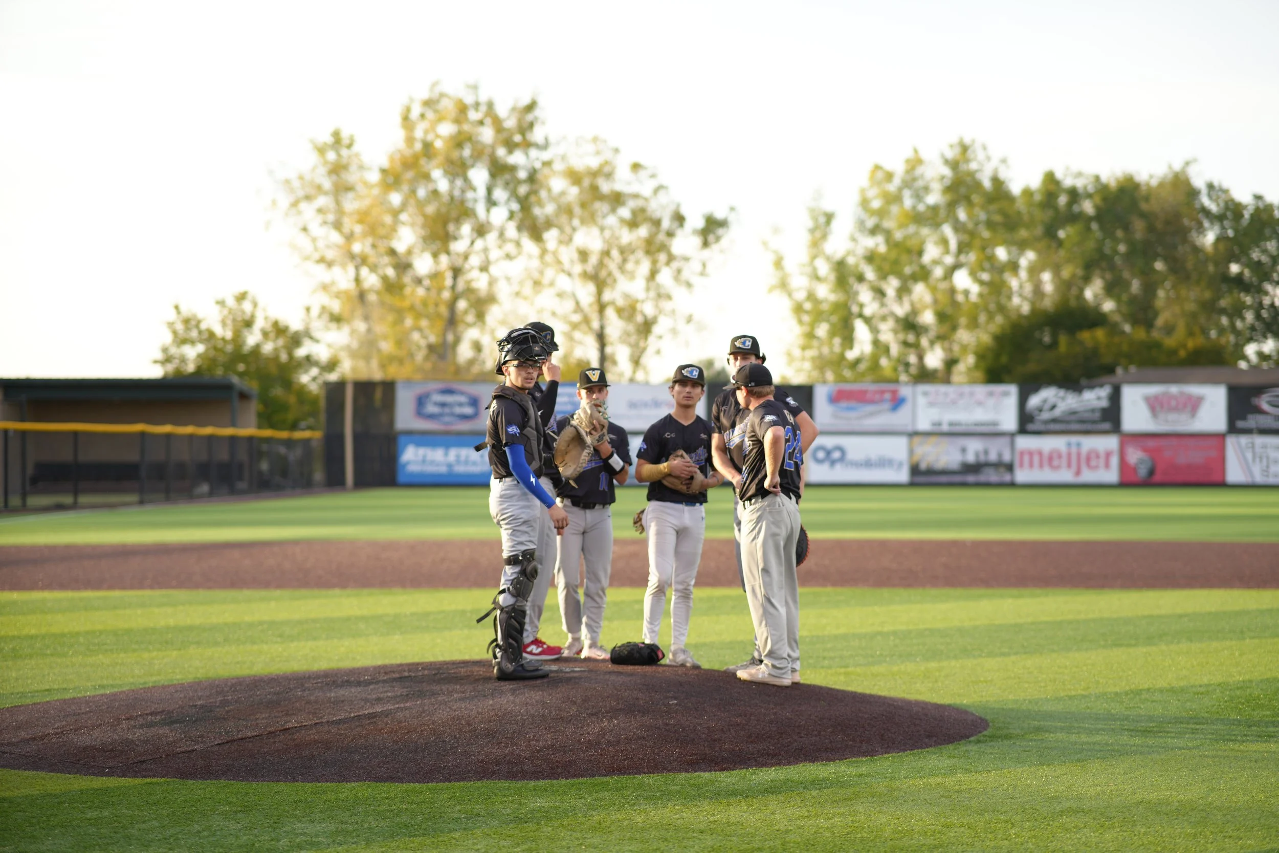 A group of five baseball players wearing black and gray uniforms and caps, standing on the pitcher's mound and talking. One player is wearing a catcher's gear. The baseball field has green grass and a brown dirt infield, with advertising banners on a fence in the background and trees with autumn foliage.