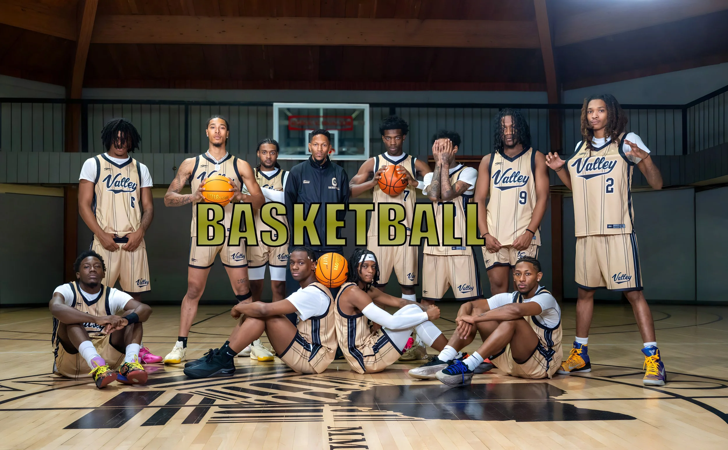 A group of basketball players in matching beige and navy uniforms on an indoor basketball court, with some sitting and others standing, holding basketballs, and posing for a team photo.