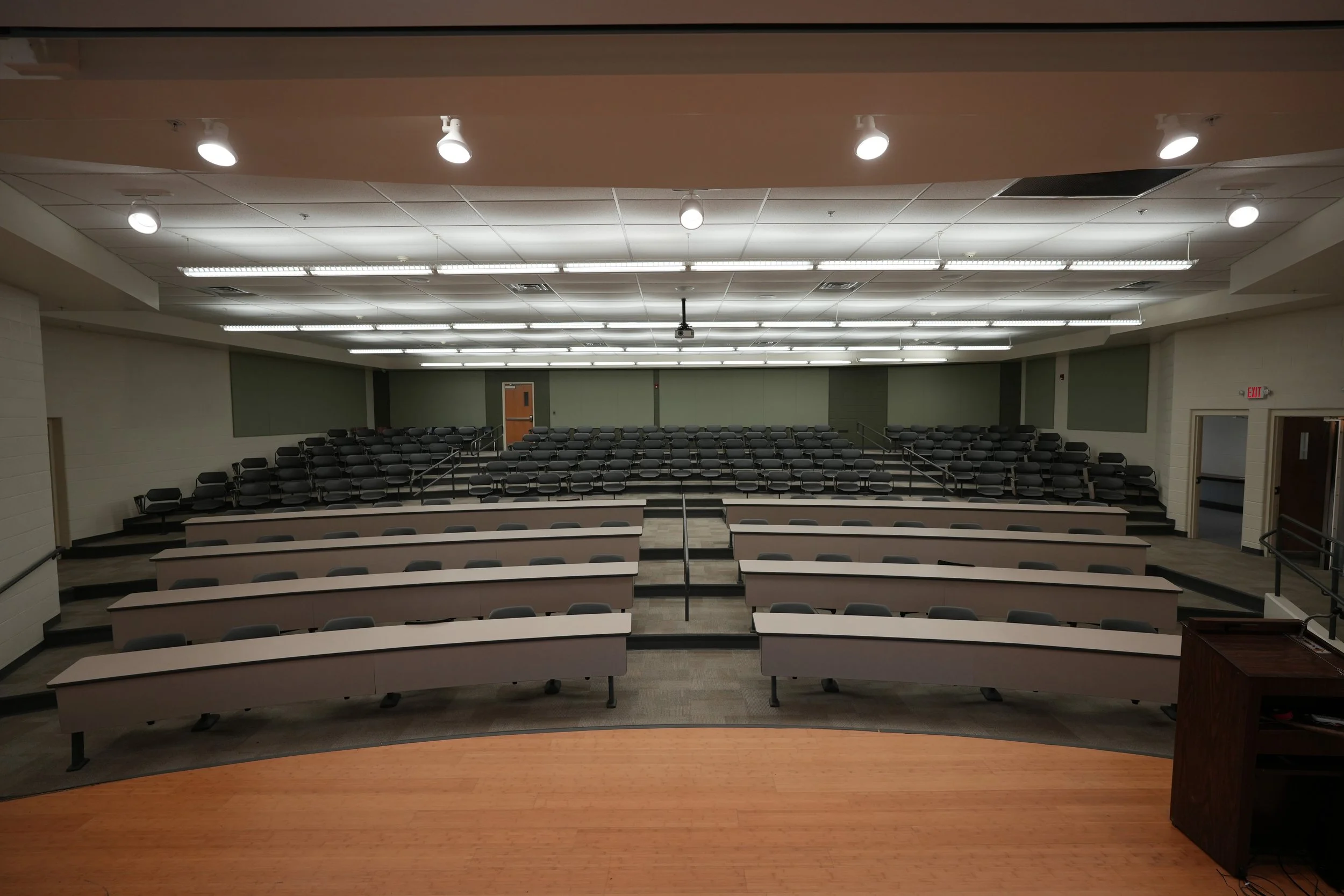 Empty university lecture hall with tiered seating, rows of desks, and a wooden stage in the foreground.