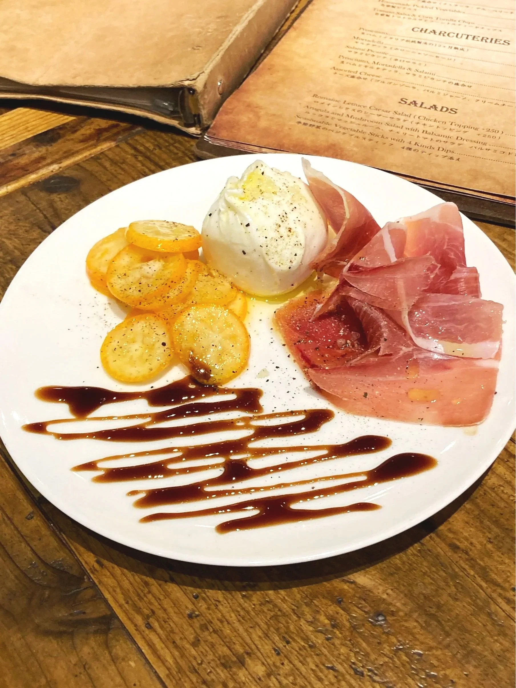 ポテト、温泉卵、パンチェッタ、バルサミコ酢ソースが盛り付けられた白い皿料理。背景にメニューブックがある。
