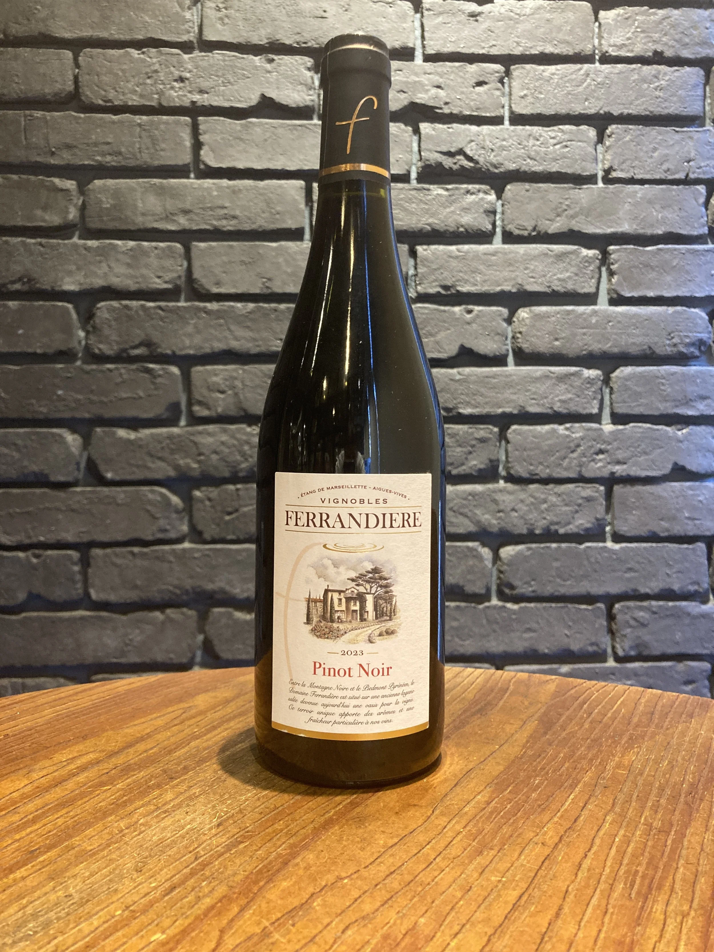 黒いラベルのワイン瓶が木製のテーブルに置かれ、背景は灰色のレンガ壁です。ラベルにはフェランディエールのワインと書かれ、ピノ・ノワールのヴィンテージ2023年と記載されています。