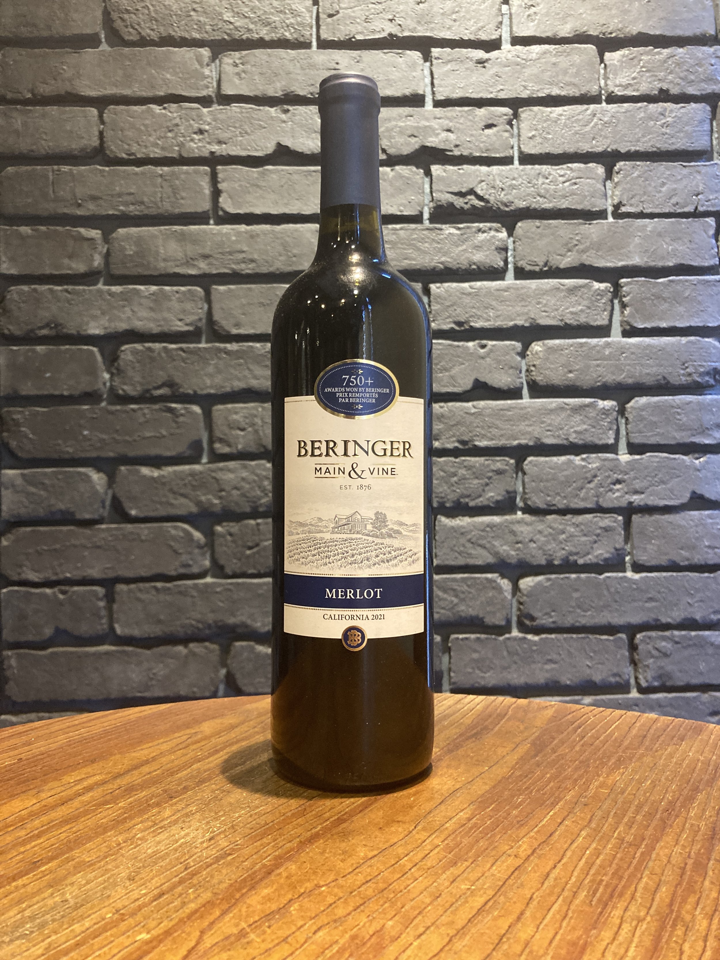 ワインのボトル、ラベルには『BERINGER MAIN & VINE』、品種はメルロー、 カリフォルニア州、2021年のヴィンテージと記載されている。背景は灰色のレンガの壁。