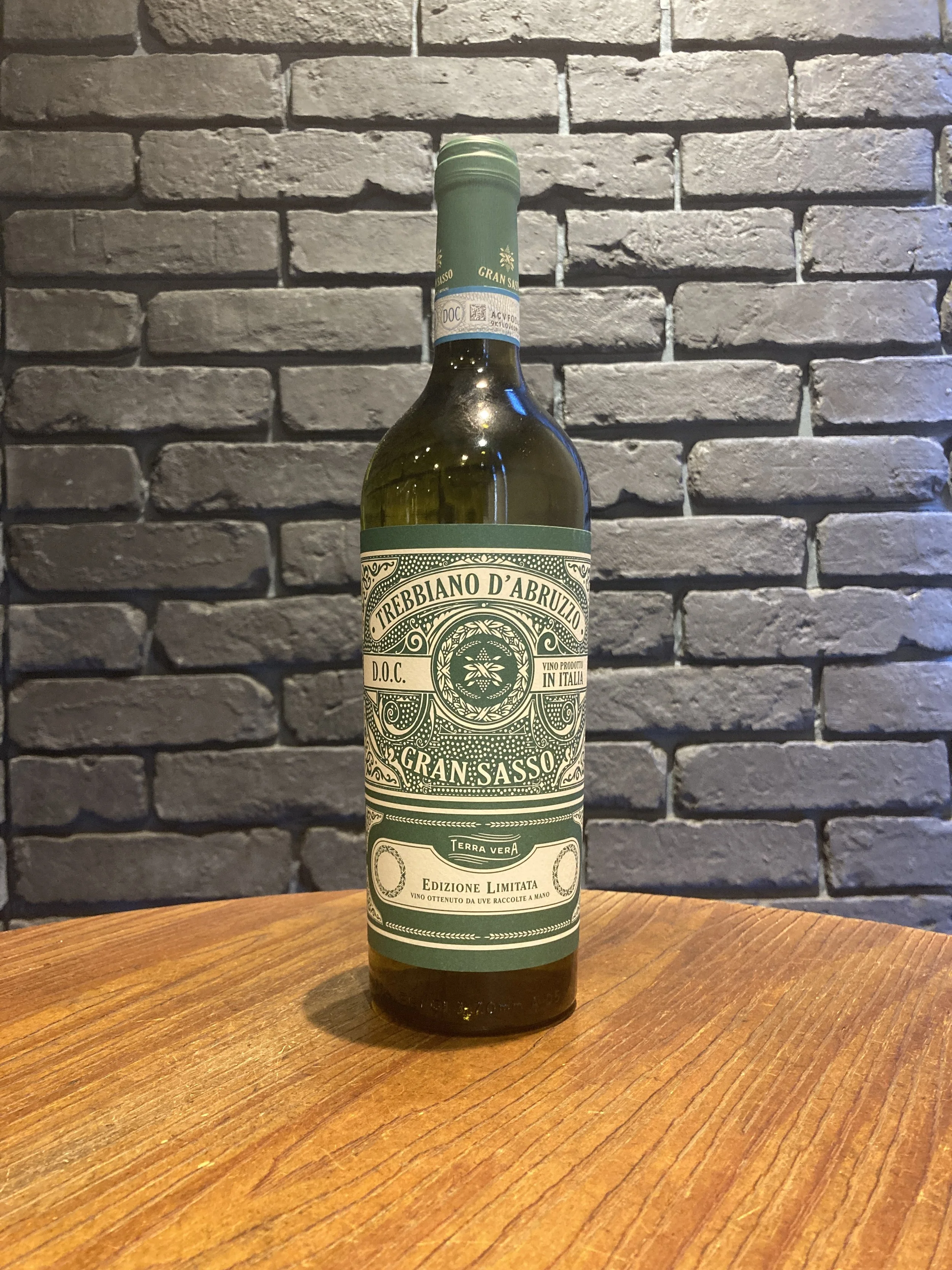 グリーンのラベルのワインボトルが木製のテーブルに置かれ、背景は灰色のレンガ壁。”}rdquo;