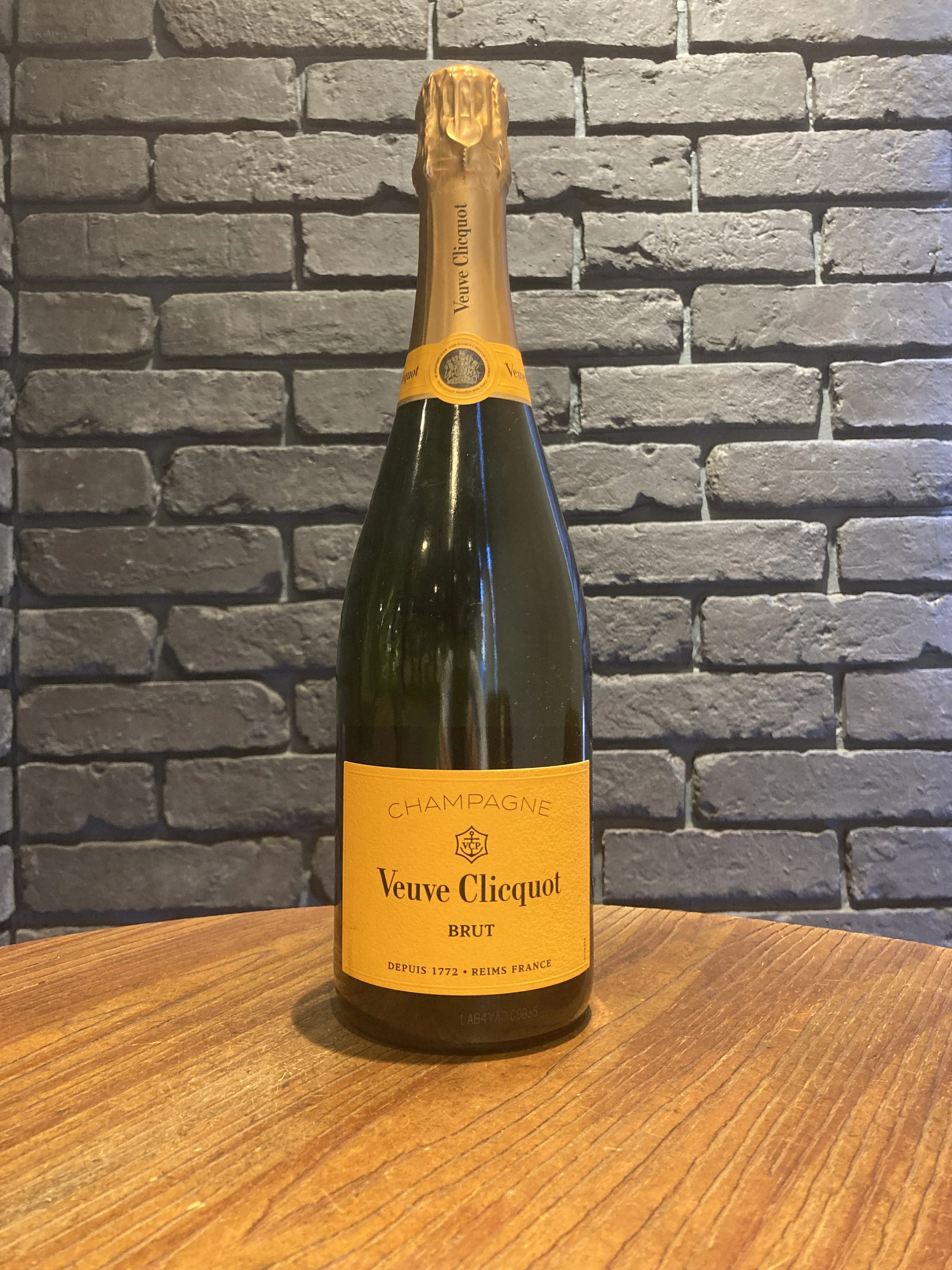 黄色いラベルのヴーヴ・クリコのスパークリングワインのボトル。ボトルは緑色で、金色のキャップがついている。背景は灰色のレンガの壁と木製のテーブルの上に置かれている。
