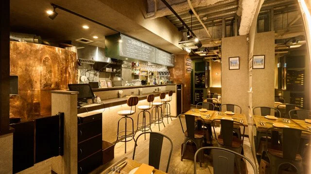 暖色照明のカフェまたはレストランの interior。カウンター席とテーブル席があり、木製のテーブルに皿とカトラリーがセットされている。カウンターにはバースツールが並び、背後にはメニューと棚に飲み物や設備が見える。