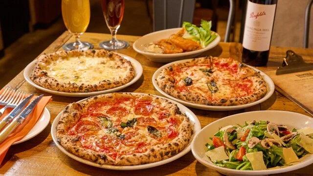 ピザ、サラダ、チキンウイング、フルーツ入りの飲み物、赤ワインがテーブルに置かれているイタリアンレストランの食事風景。
