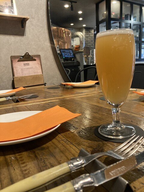 木製のテーブルにグラスに入ったビール、白い皿とオレンジのナプキン、カトラリーが置かれている。背景にカウンターと調理設備が見える飲食店の interior.