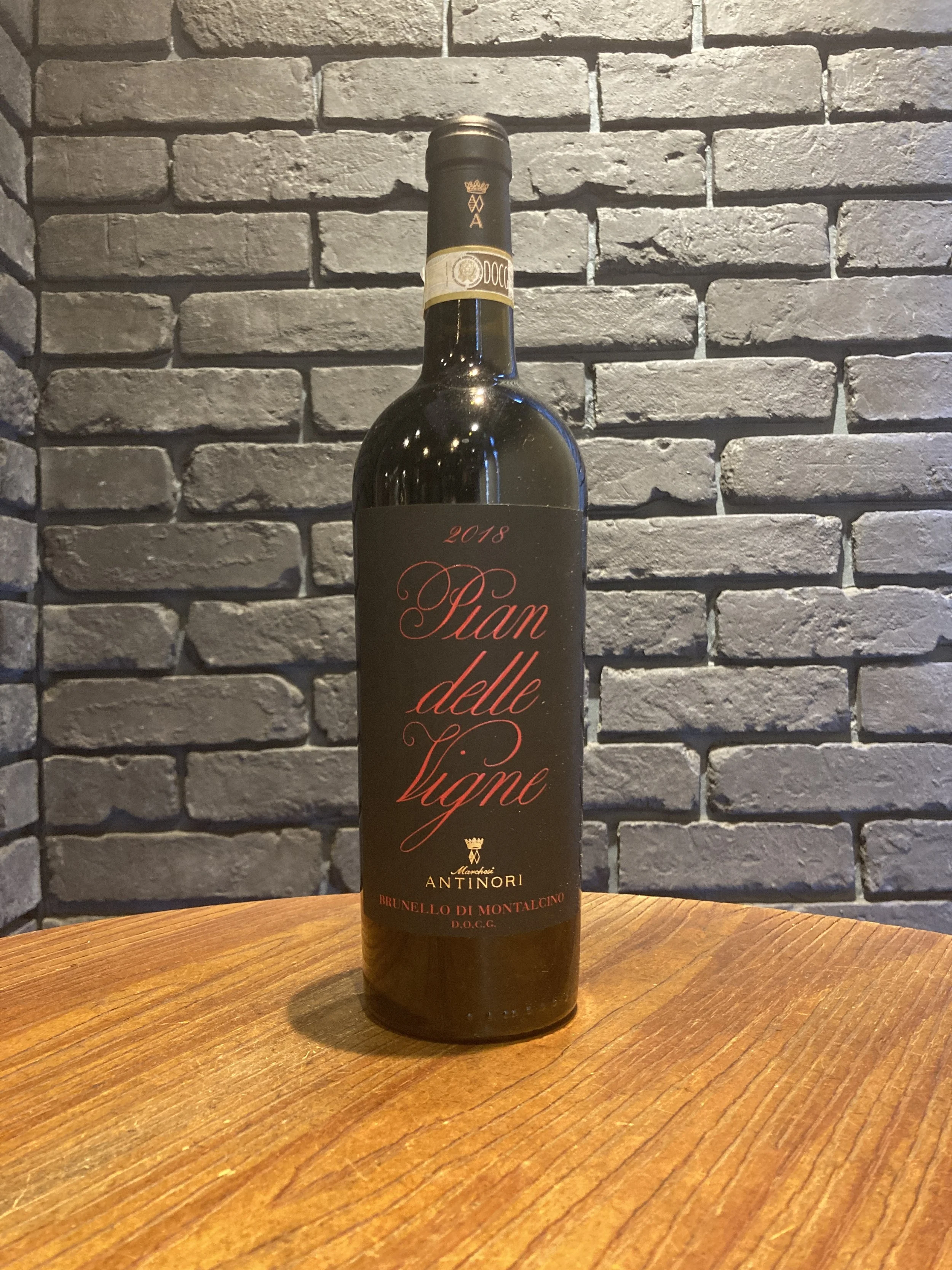 黒いラベルの赤い文字のワインボトルが木製のテーブルに立っている。背景は灰色のレンガの壁。