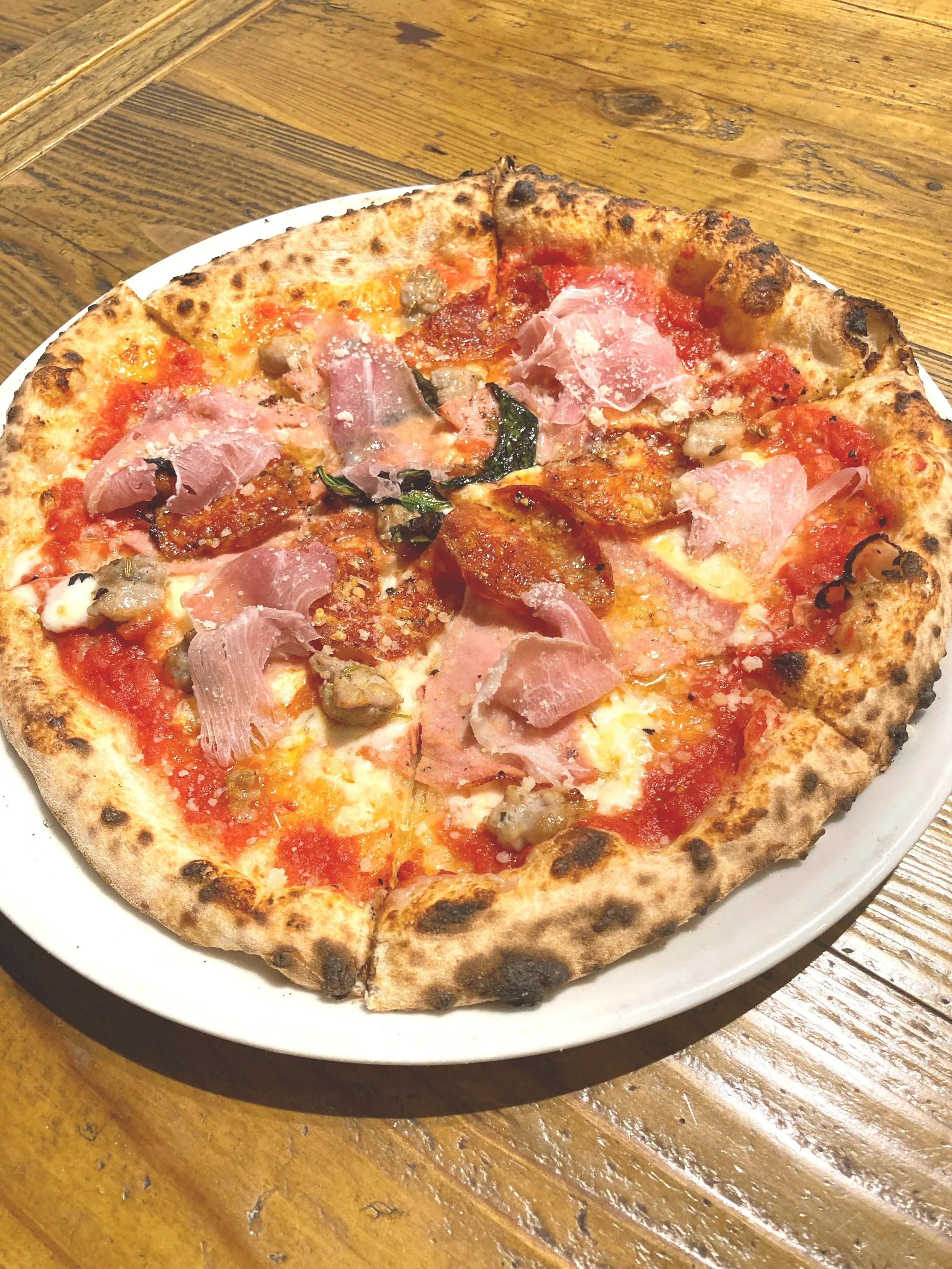 ピザが皿に乗っている。トマトソース、チーズ、ハム、サラミ、野菜が乗っている。