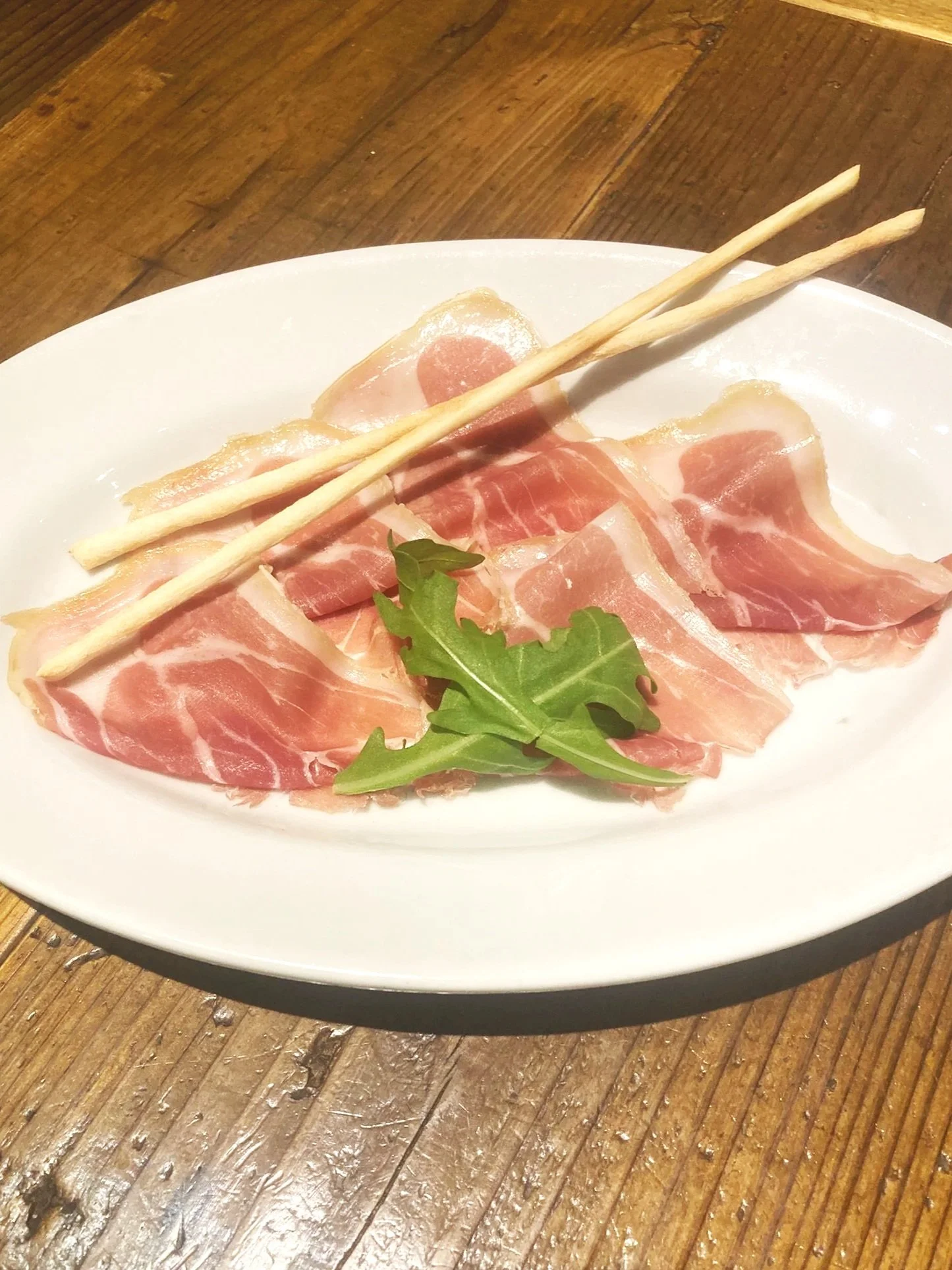 生ハムと葉っぱが盛り付けられた白い楕円形の皿の上に、棒状のクラッカーが2本置かれている。