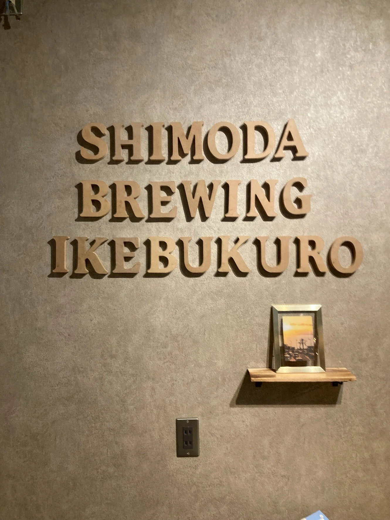壁に木製の看板で、「SHIMODA BREWING IKEBUKURO」と書かれている。下に小さな棚と写真フレームが置かれている壁の一部分。
