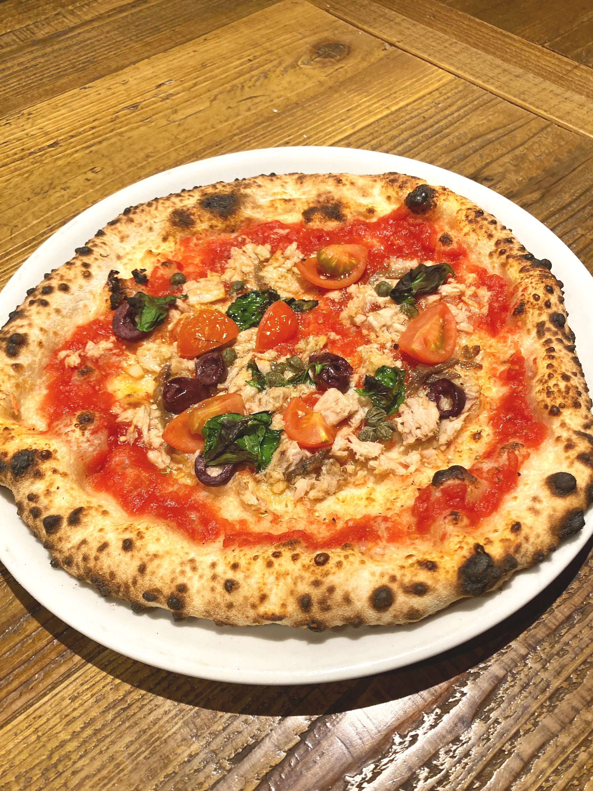 木製のテーブルの上に置かれた白い皿に乗ったピザ。焼き色のついたクラストとトマトソース、トマト、黒オリーブ、鶏肉、バジルが乗っている。