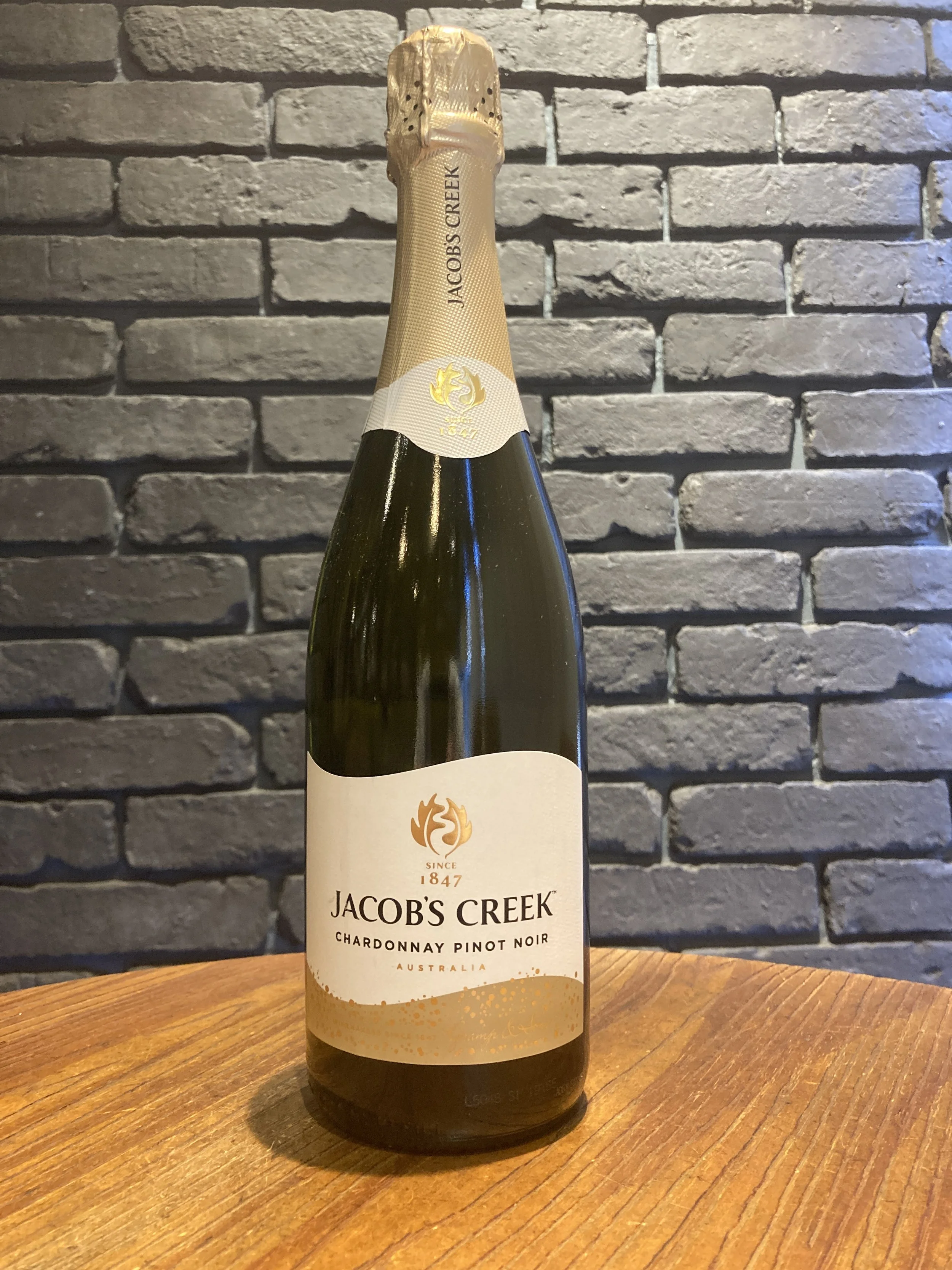 木製のテーブルの上に置かれたスパークリングワインのボトル。背景は灰色のレンガ壁で、ラベルには"JACOB'S CREEK"と書かれている。