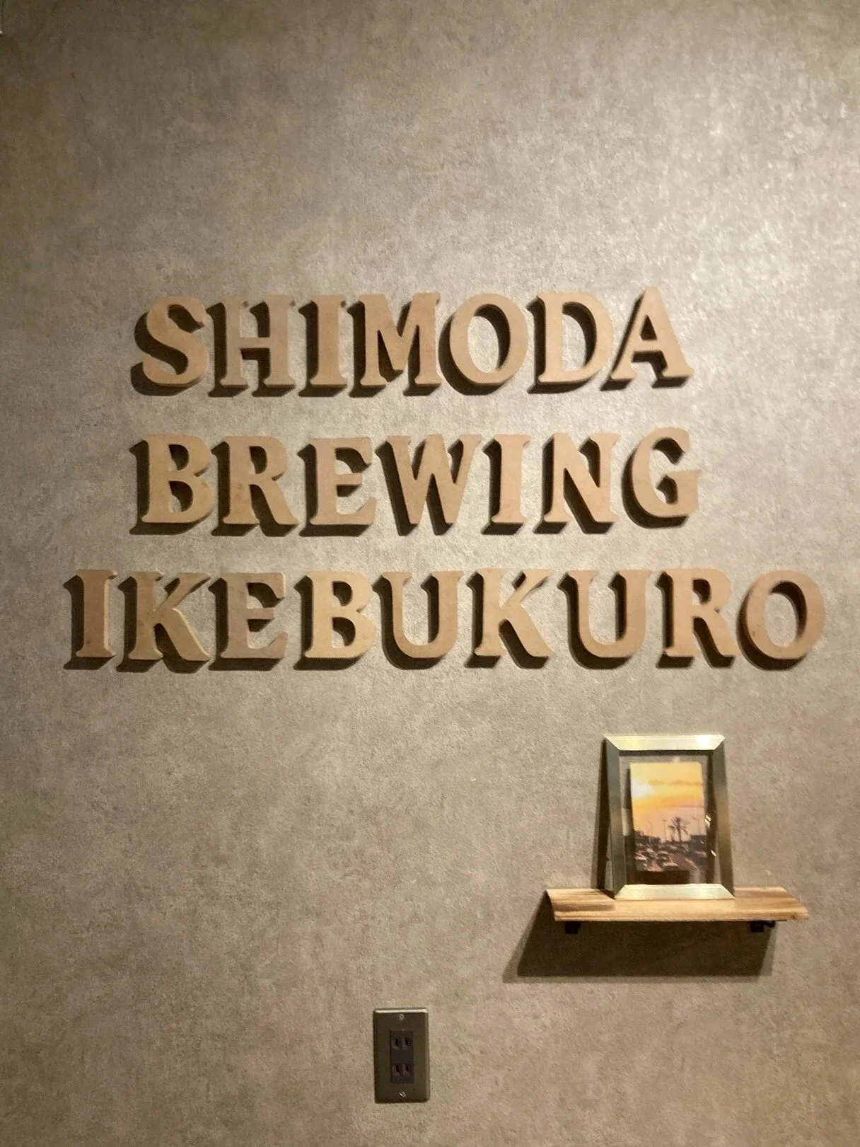 壁に木の文字で書かれた看板。SHIMODA BREWING IKEBUKUROと書かれている。下には小さな棚と写真フレームがある。」}