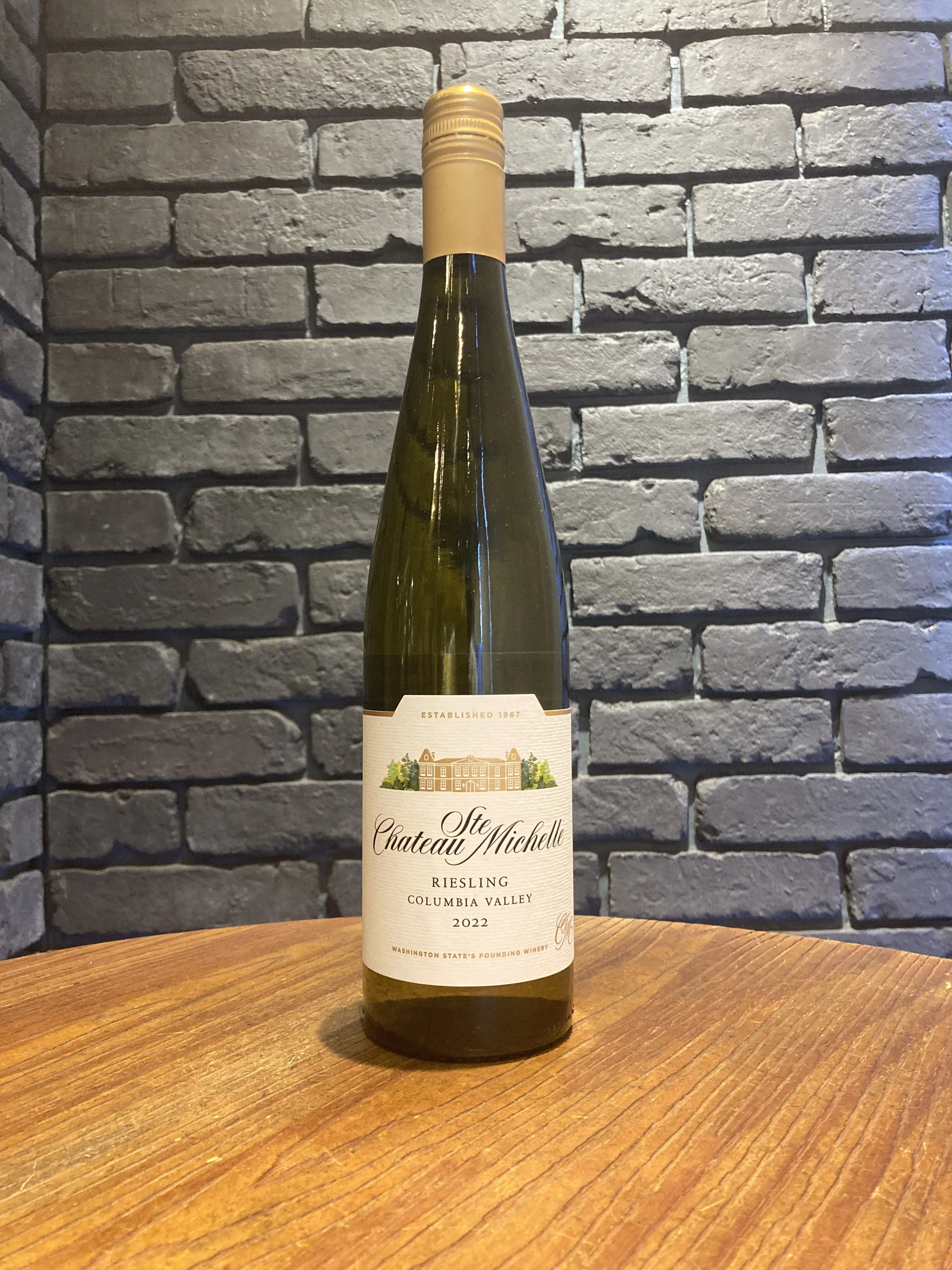 コルビージャングルズの白ワインのボトルが木製のテーブルに置かれ、背後に黒いレンガの壁が映る。