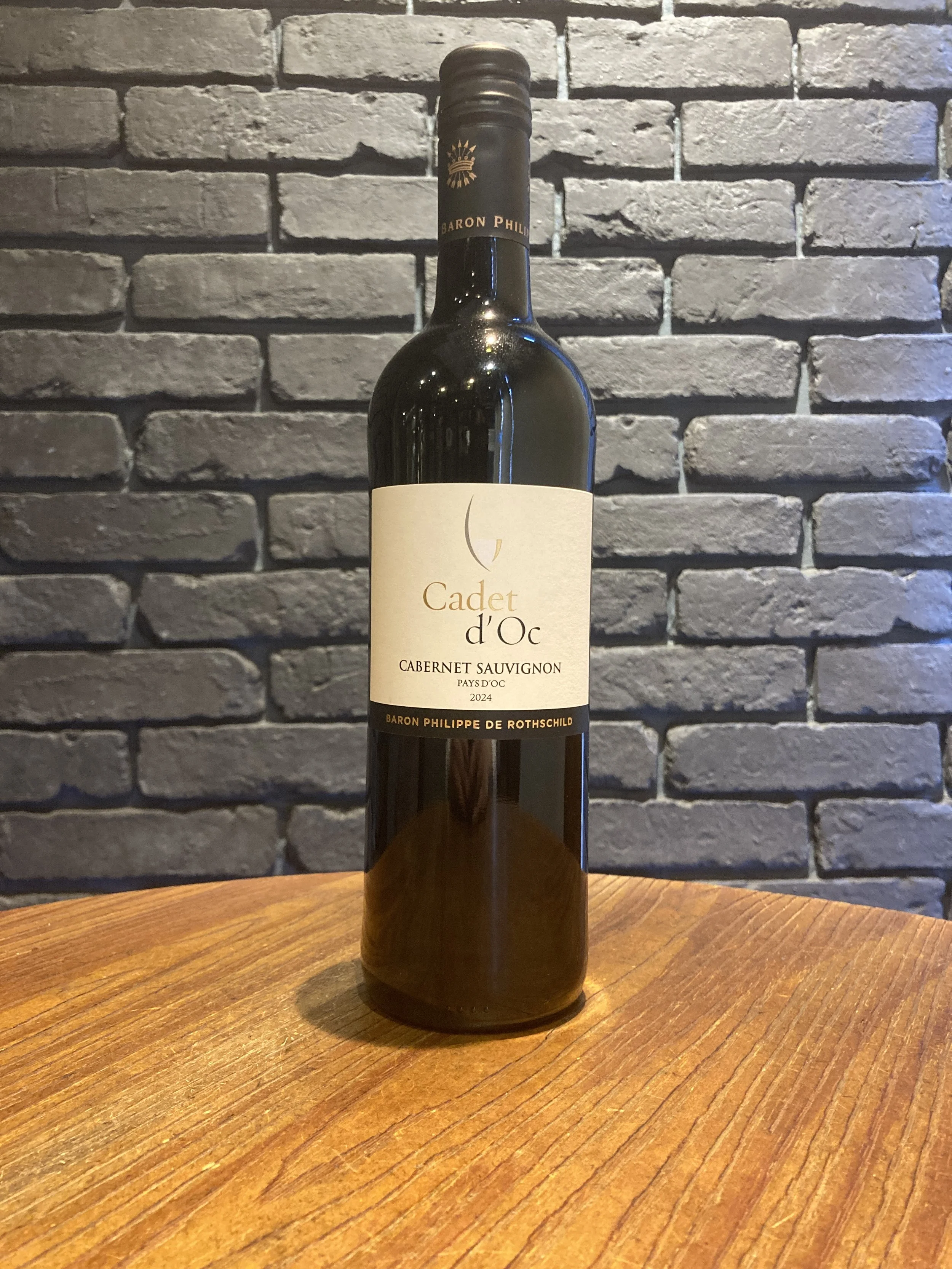 黒いラベルのワインボトルが木製のテーブルの上に立っており、背景は灰色のレンガ壁です。